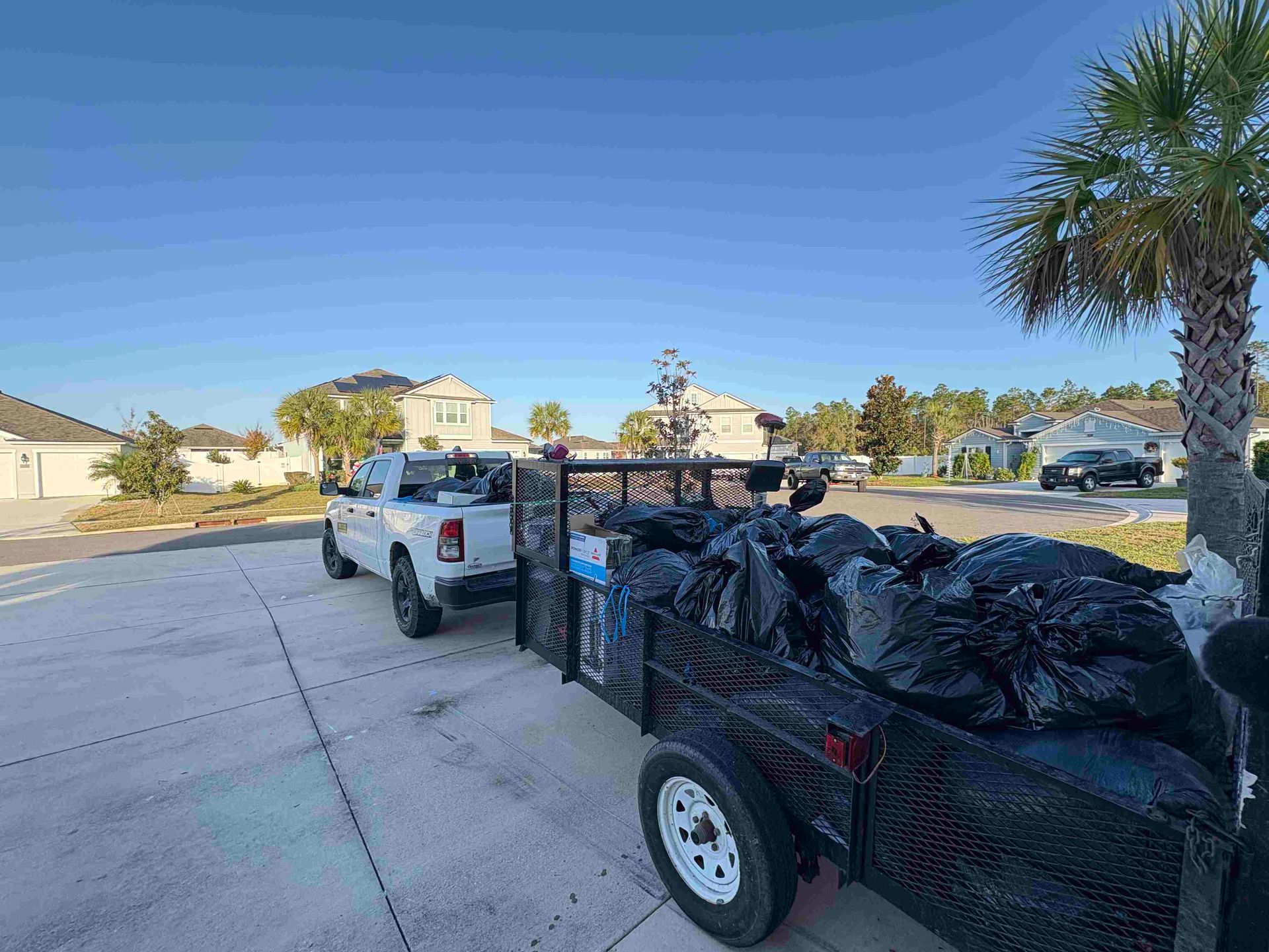 Truck with trailer loaded with black trash bags on a paved driveway in front of suburban houses.