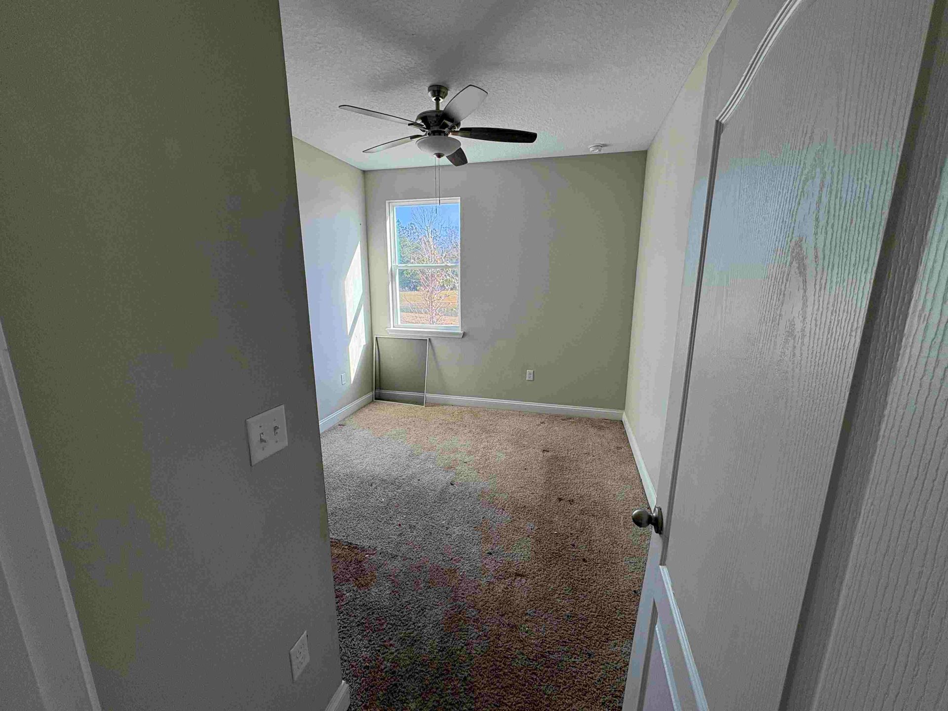 Empty bedroom with tan carpet, a window, and a ceiling fan.