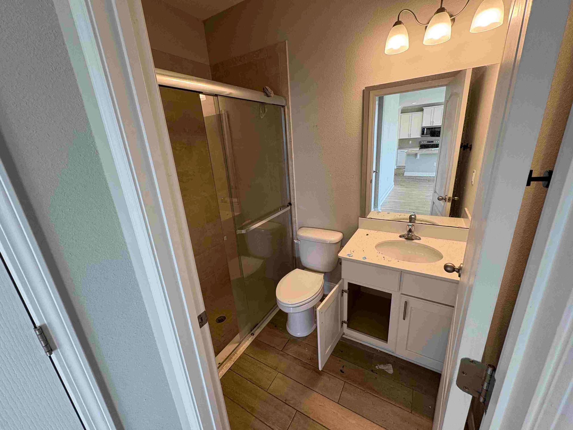 Bathroom with a toilet, shower, vanity, and a partially open door. Beige walls, wood-look flooring.