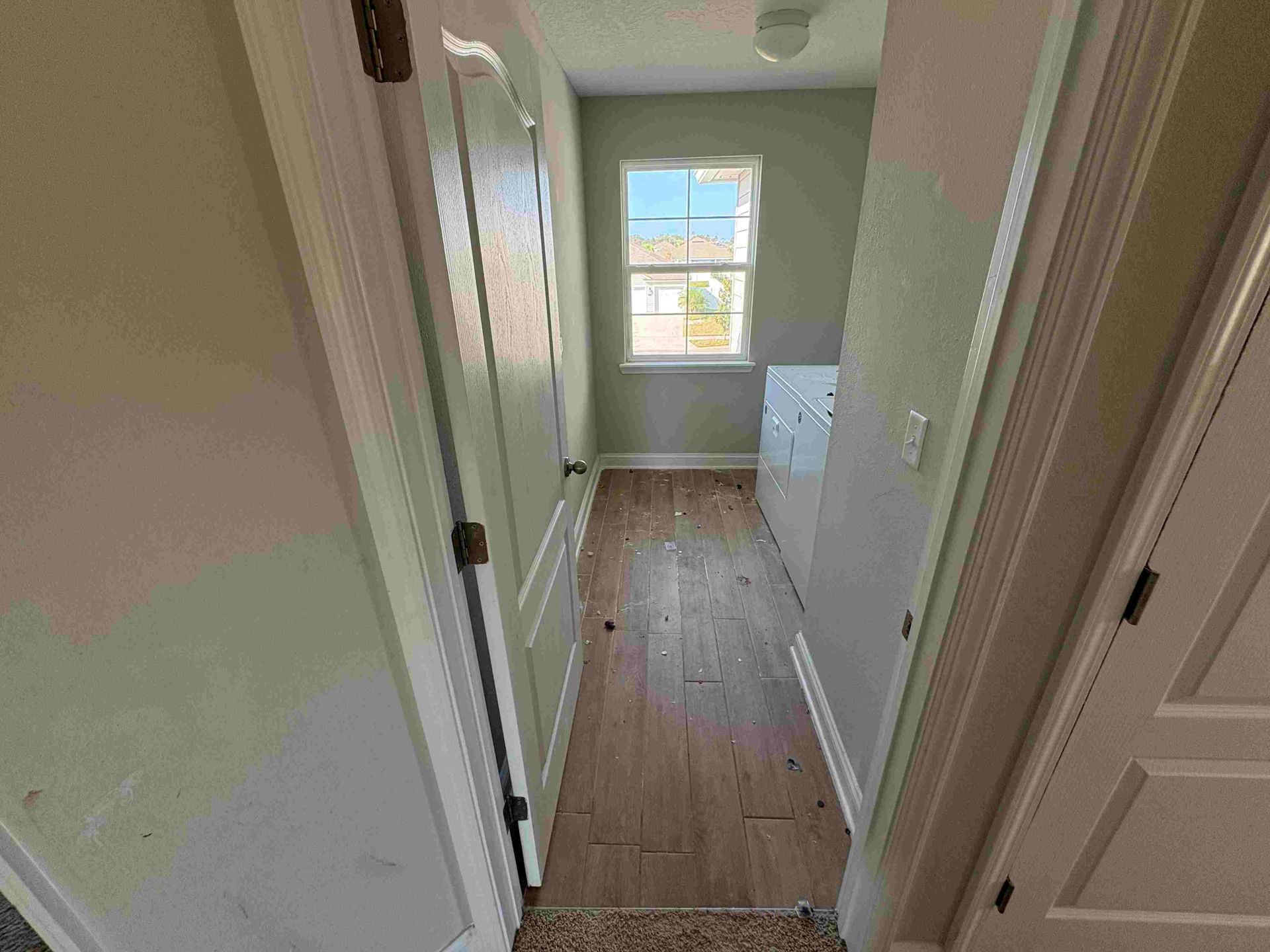 Narrow hallway with wooden floor, white doors, and window at the end, leading to a utility room.