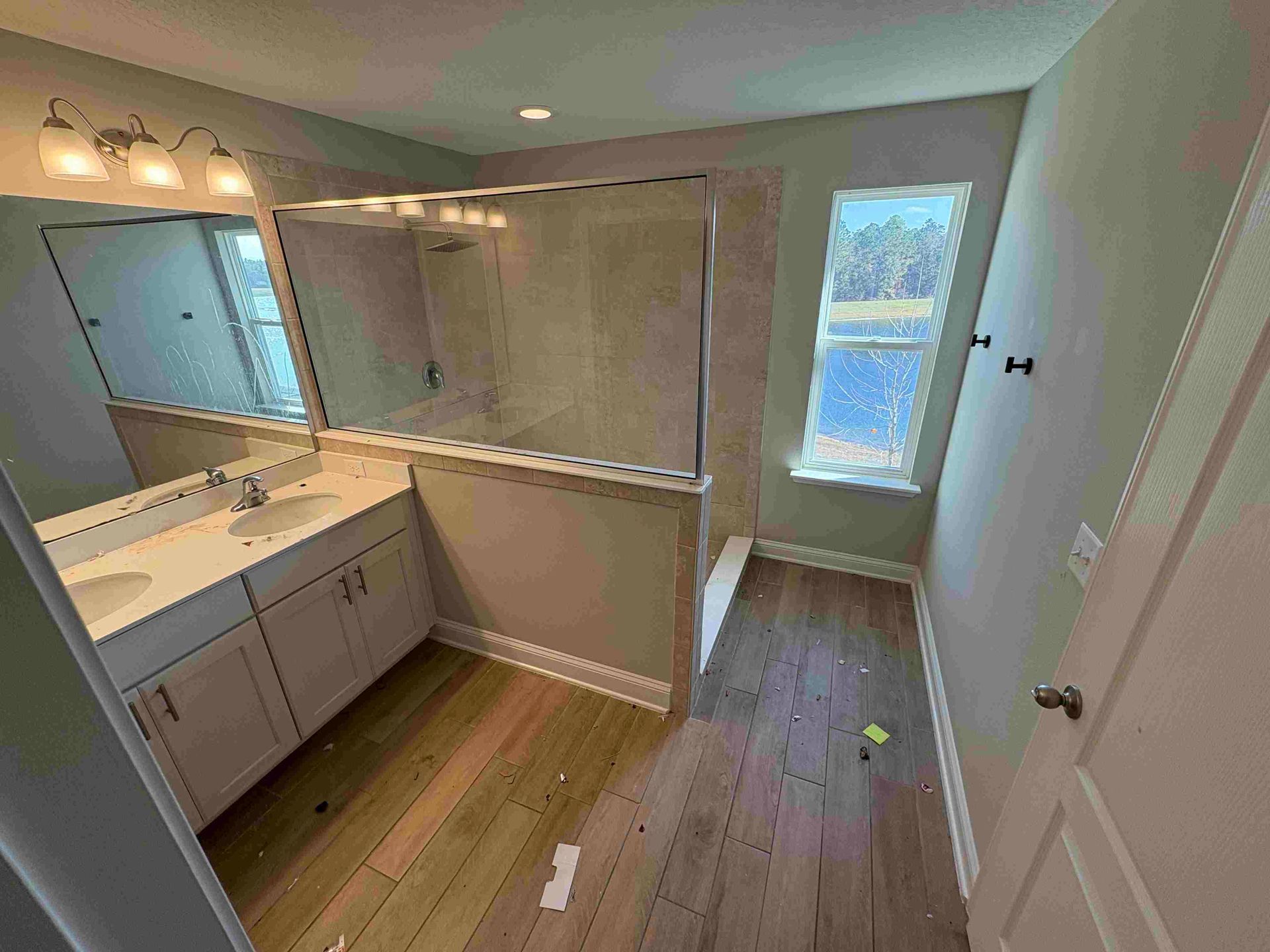 Bathroom with a double sink vanity, shower, and window. Beige walls and wooden floors.