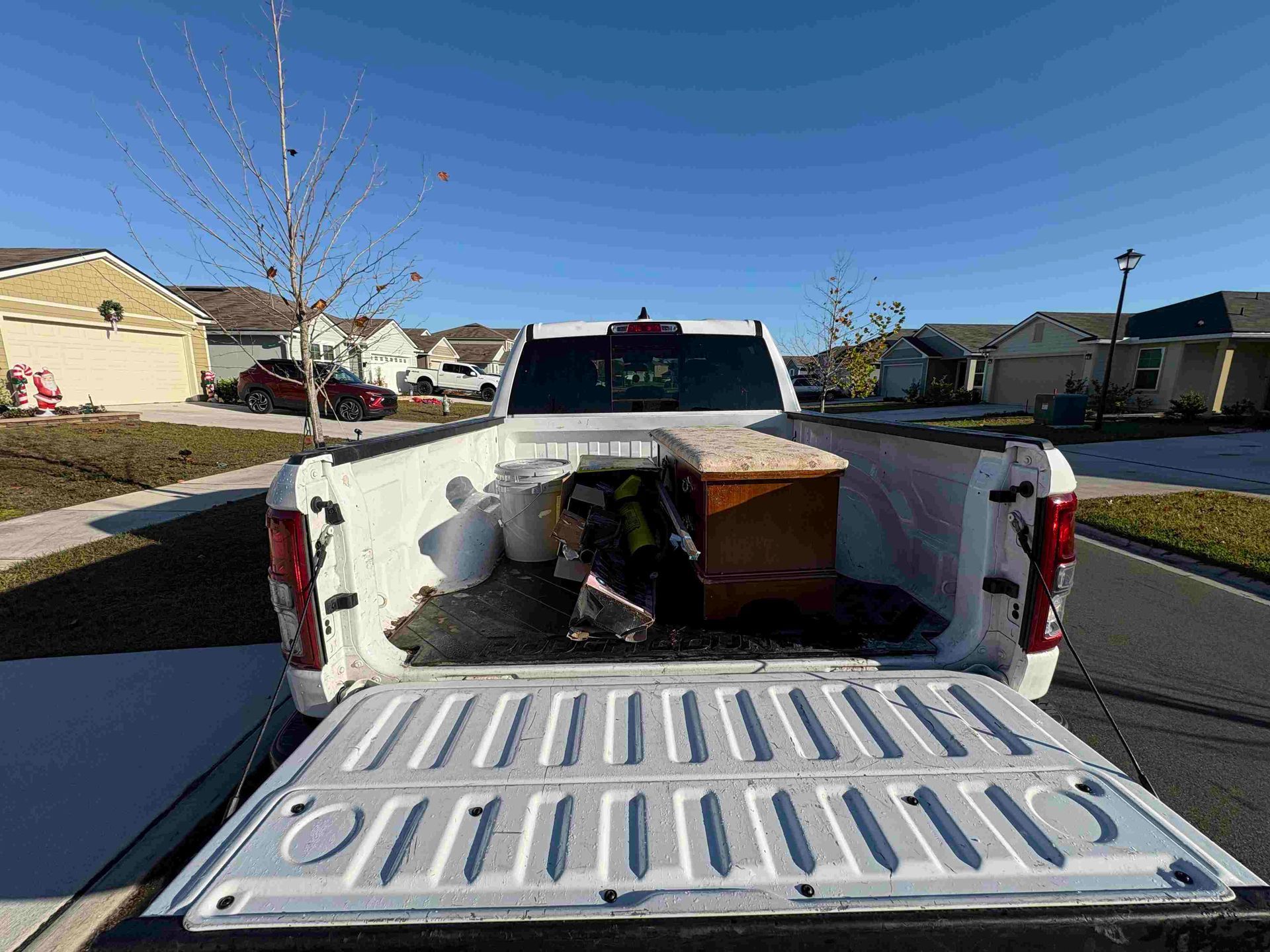 White pickup truck bed open, containing items: wooden box, bags, debris. Sunny residential street background.