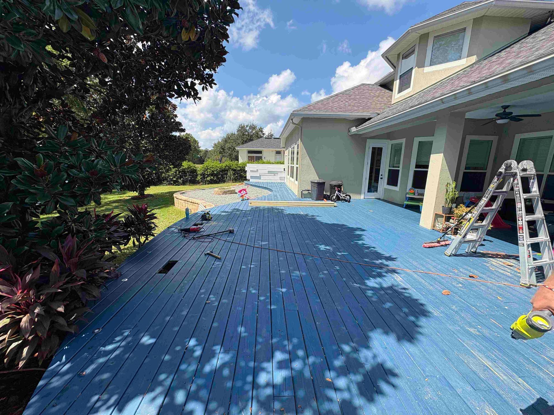 Blue-painted wooden deck partially dismantled near a two-story house under a sunny sky. Construction tools and a ladder visible.