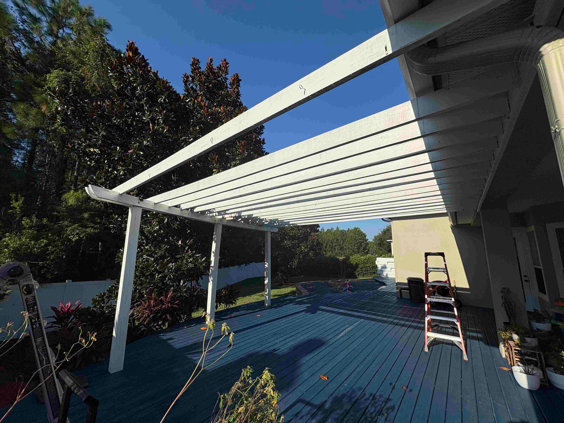White pergola over a blue deck, surrounded by trees and a house under a clear sky.