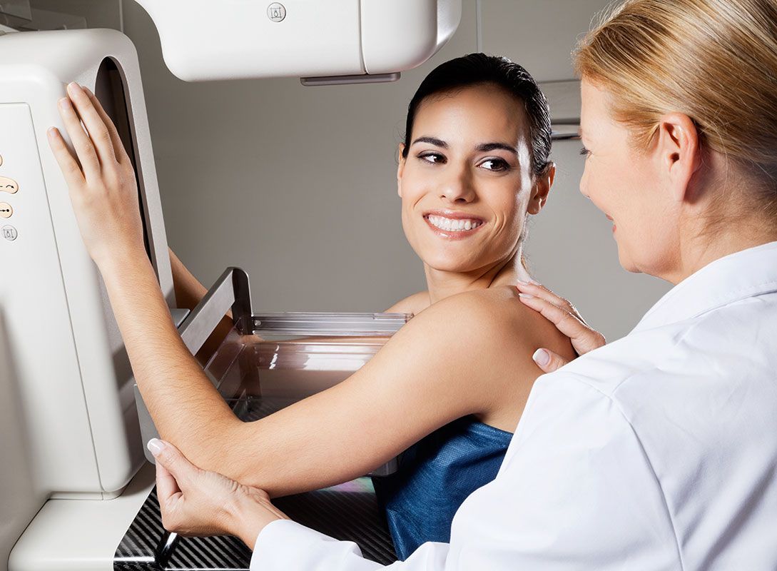 Woman smiling during a mammogram; healthcare worker assists.