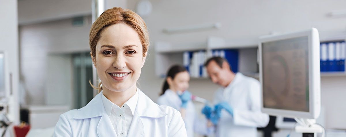 Woman in lab coat smiles at the camera in a lab setting, with colleagues in background.