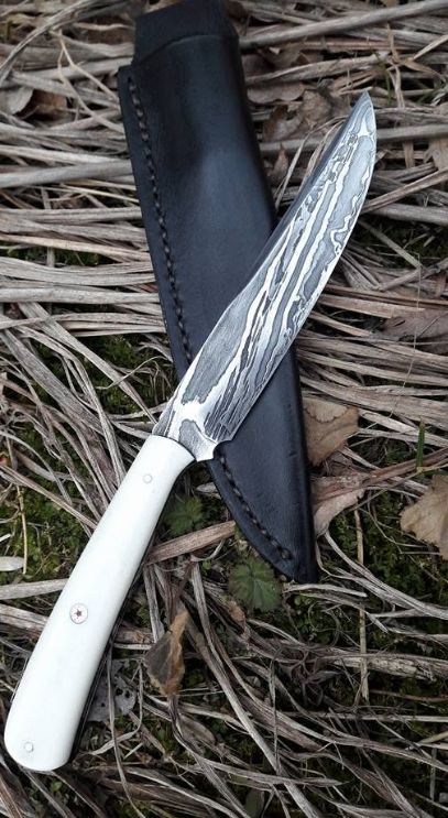 White-handled knife with patterned blade in black sheath on grass.