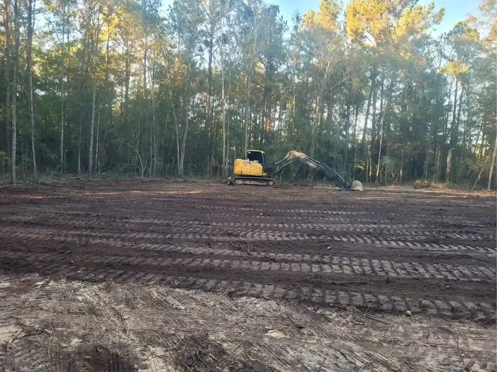 A yellow excavator parked on a cleared, muddy lot with tire tracks, positioned before a dense treeline.
