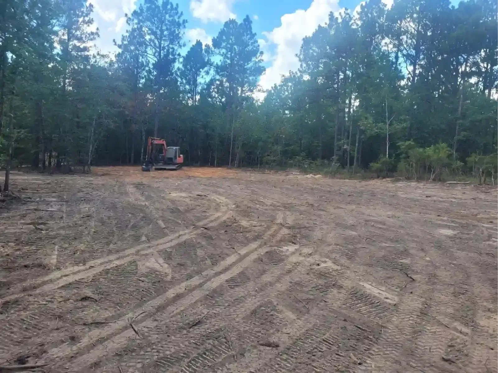 A cleared dirt lot with tire tracks, featuring a small excavator in the background near a dense line of pine trees.