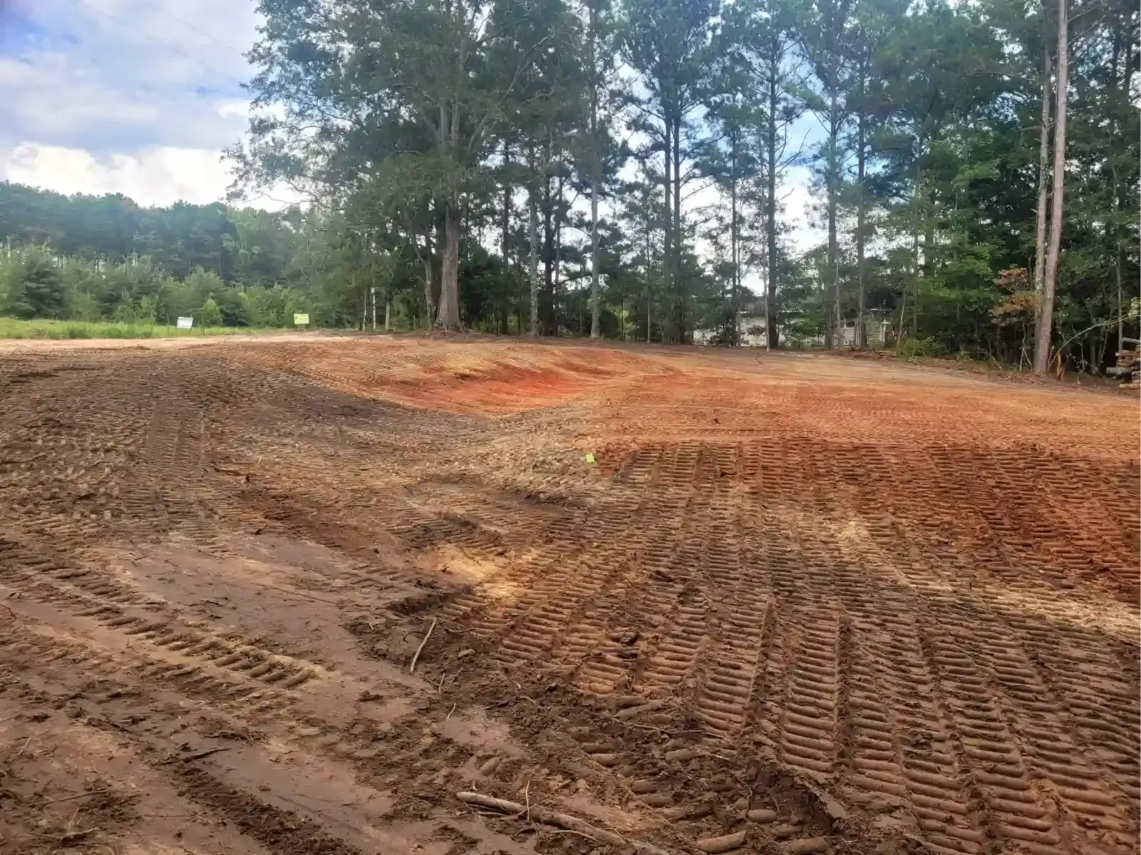 A cleared lot with exposed red and brown soil, marked by tire tracks and framed by a line of trees against a blue sky.