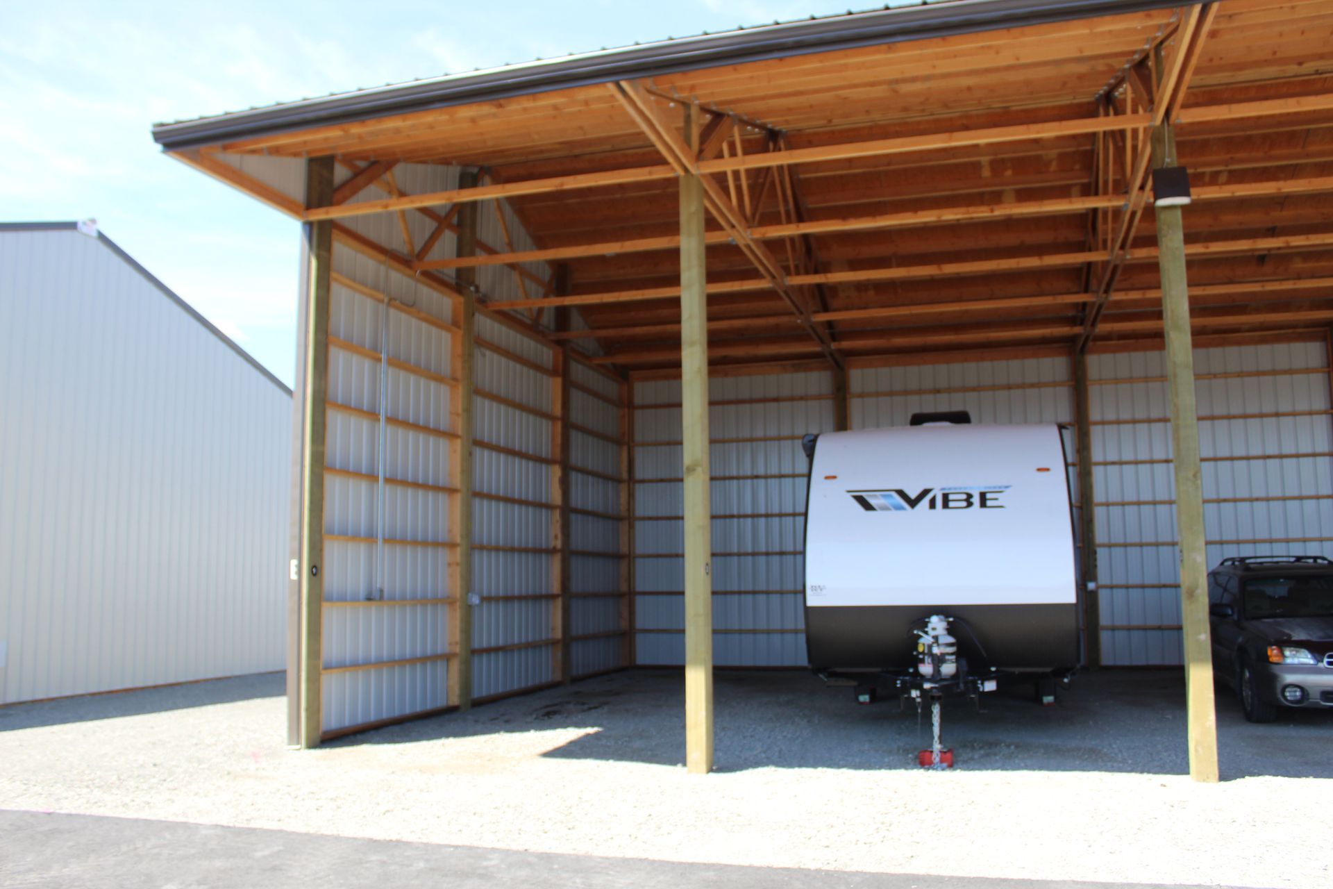 RV parked under a wooden carport; metal siding and roof; gravel ground; another building in the background.