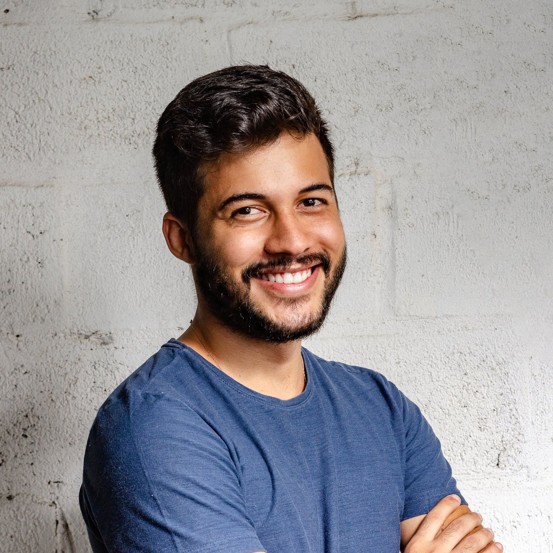 A man with a beard wearing a blue shirt is smiling with his arms crossed.