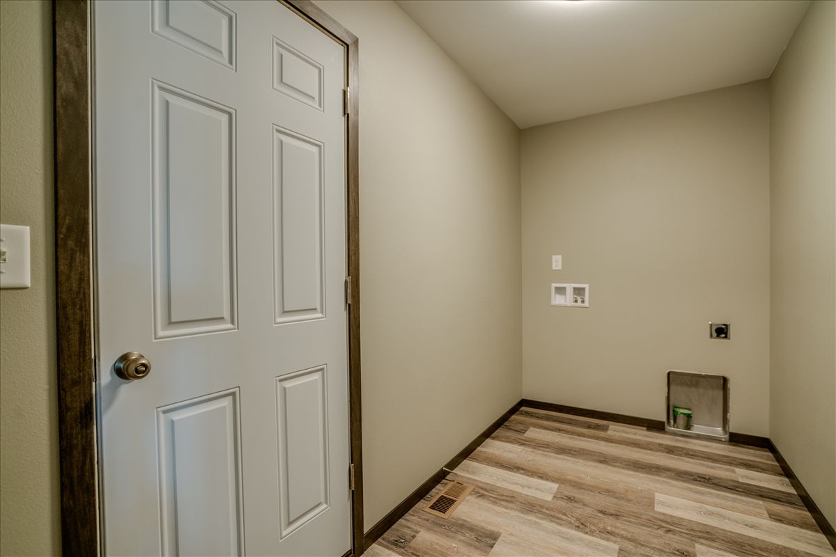 An indoor laundry hookup space with light-colored walls, wood-look flooring, and a white interior door.