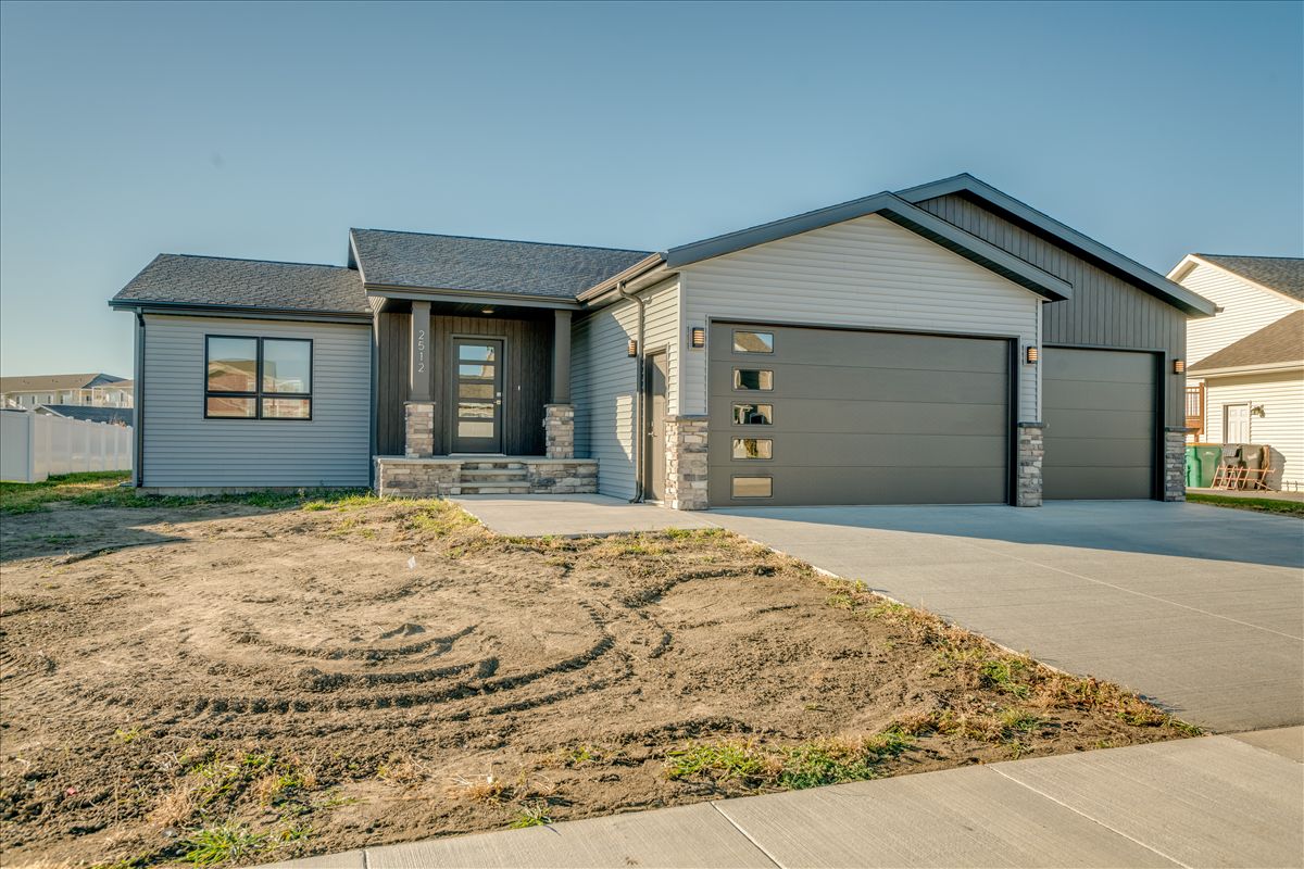 A single-story suburban house with grey siding, stone accents, and a multi-car garage, fronted by an unfinished dirt yard.