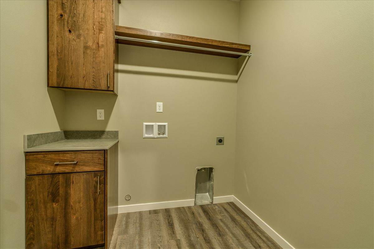 A laundry room with light beige walls, wood-finish cabinets and shelving, and gray wood-look vinyl flooring.