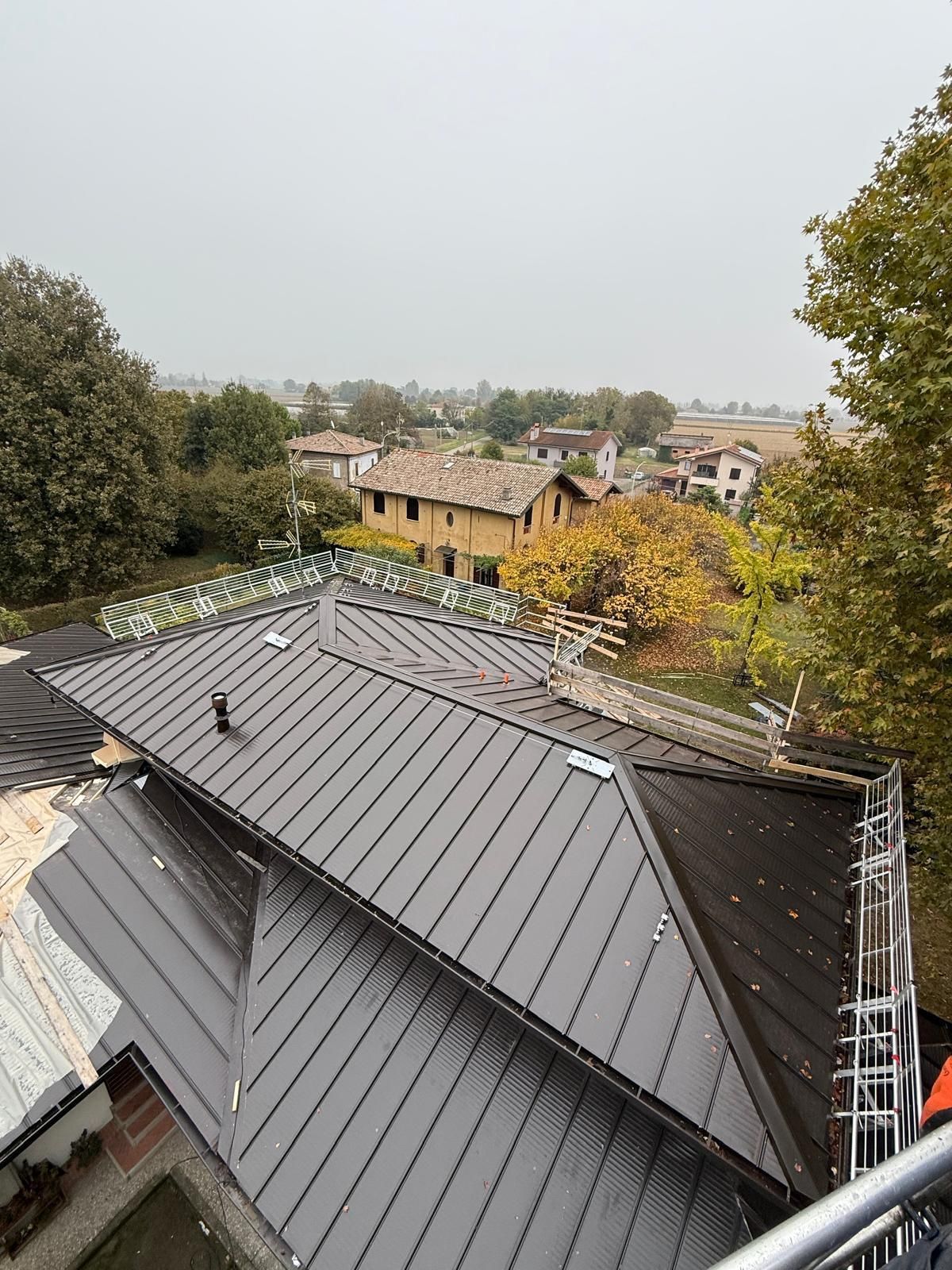 Installazione del tetto in metallo in corso, contesto rurale con impalcature e case sullo sfondo. Cielo coperto.