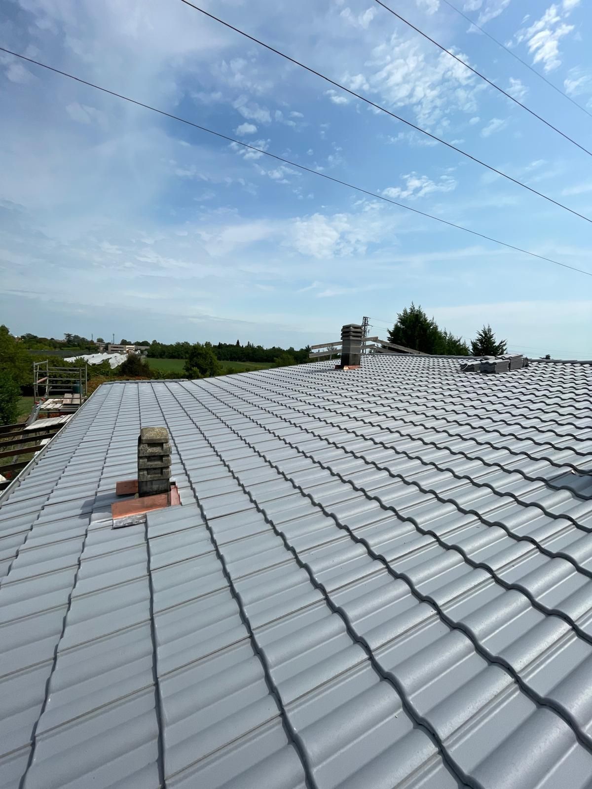Tetto in metallo grigio con due camini contro un cielo azzurro con linee elettriche.