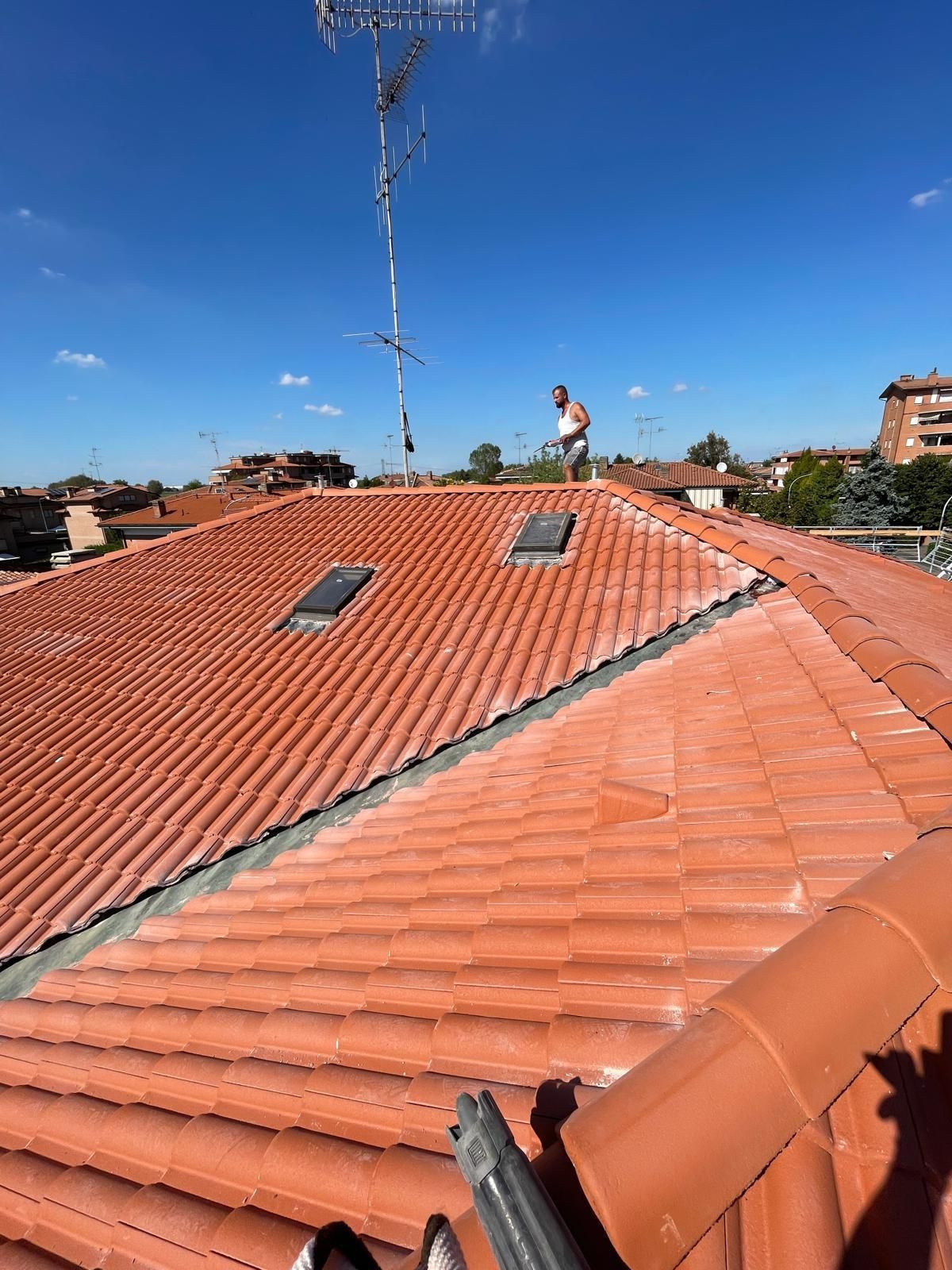 Uomo su un tetto di tegole rosse sotto il cielo azzurro, vicino all'antenna e ai lucernari, in un contesto residenziale.
