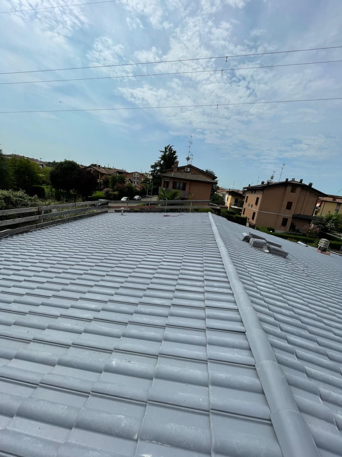 Tetto di tegole grigie con un tubo di metallo lungo il colmo, case e alberi sullo sfondo sotto un cielo nuvoloso.