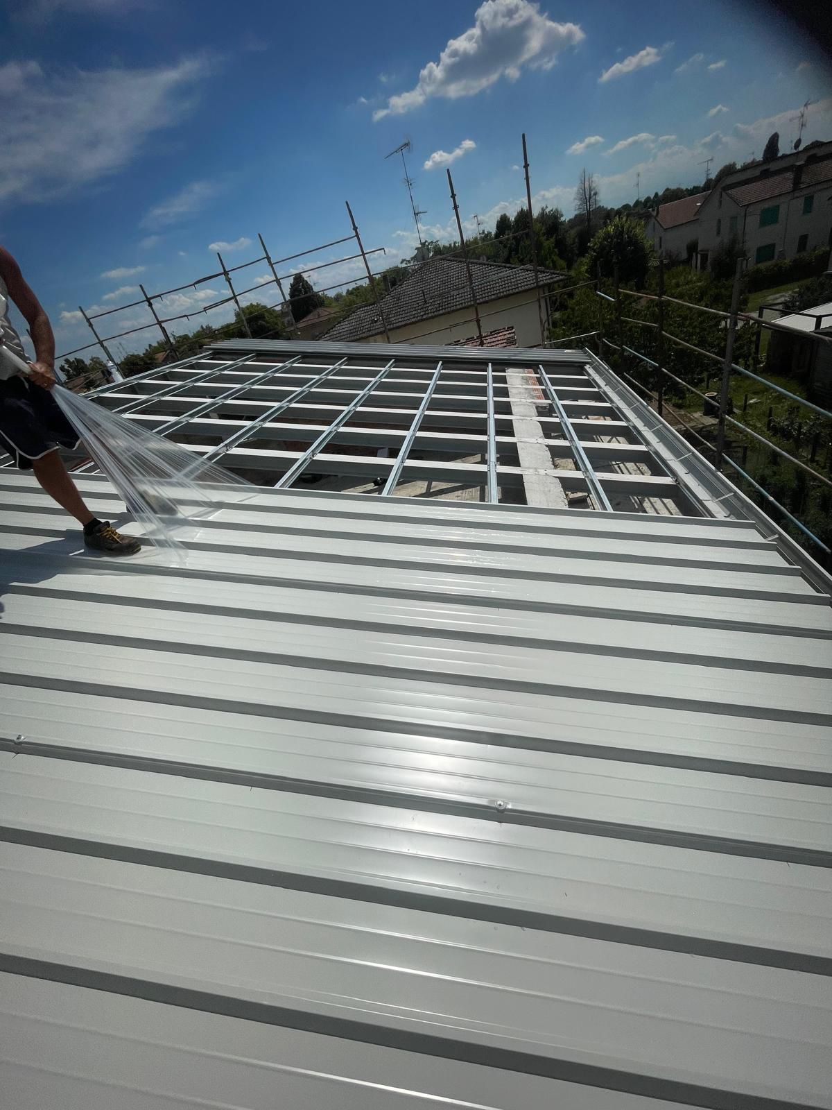 Uomo che installa una rete su un tetto in lamiera ondulata con struttura a vista e case residenziali sullo sfondo.