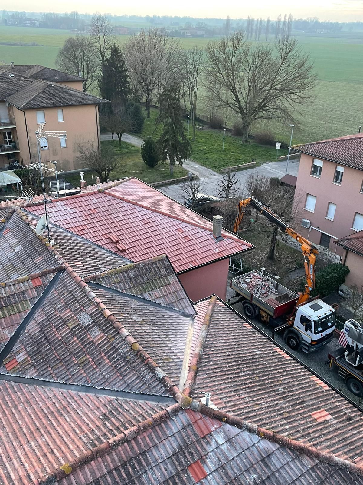 Vista dall'alto di edifici con tetti di tegole rosse, un cantiere con un escavatore e un camion.