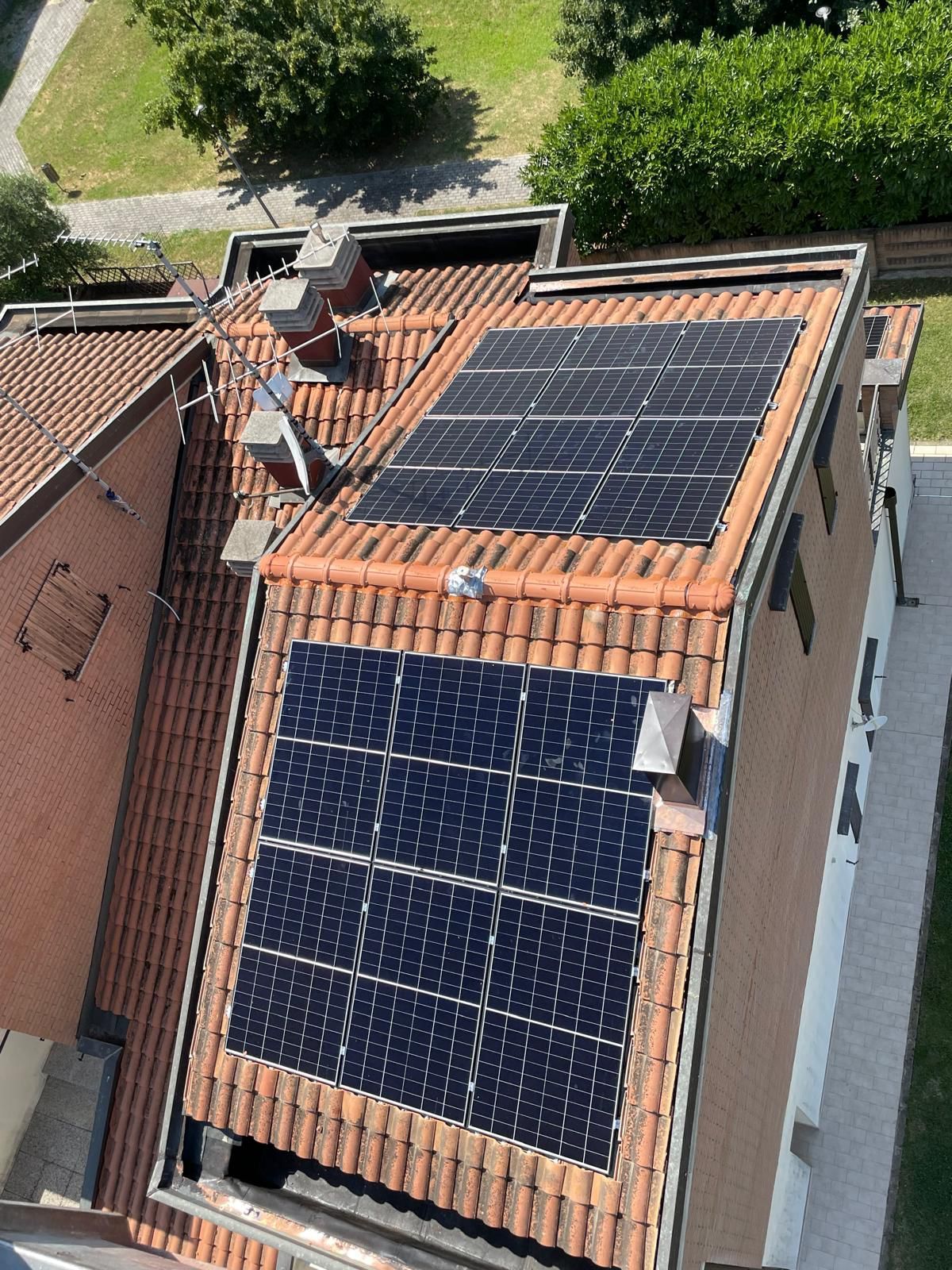 Tetto con tegole rosse e due serie di pannelli solari blu scuro. L'edificio è circondato da alberi verdi.