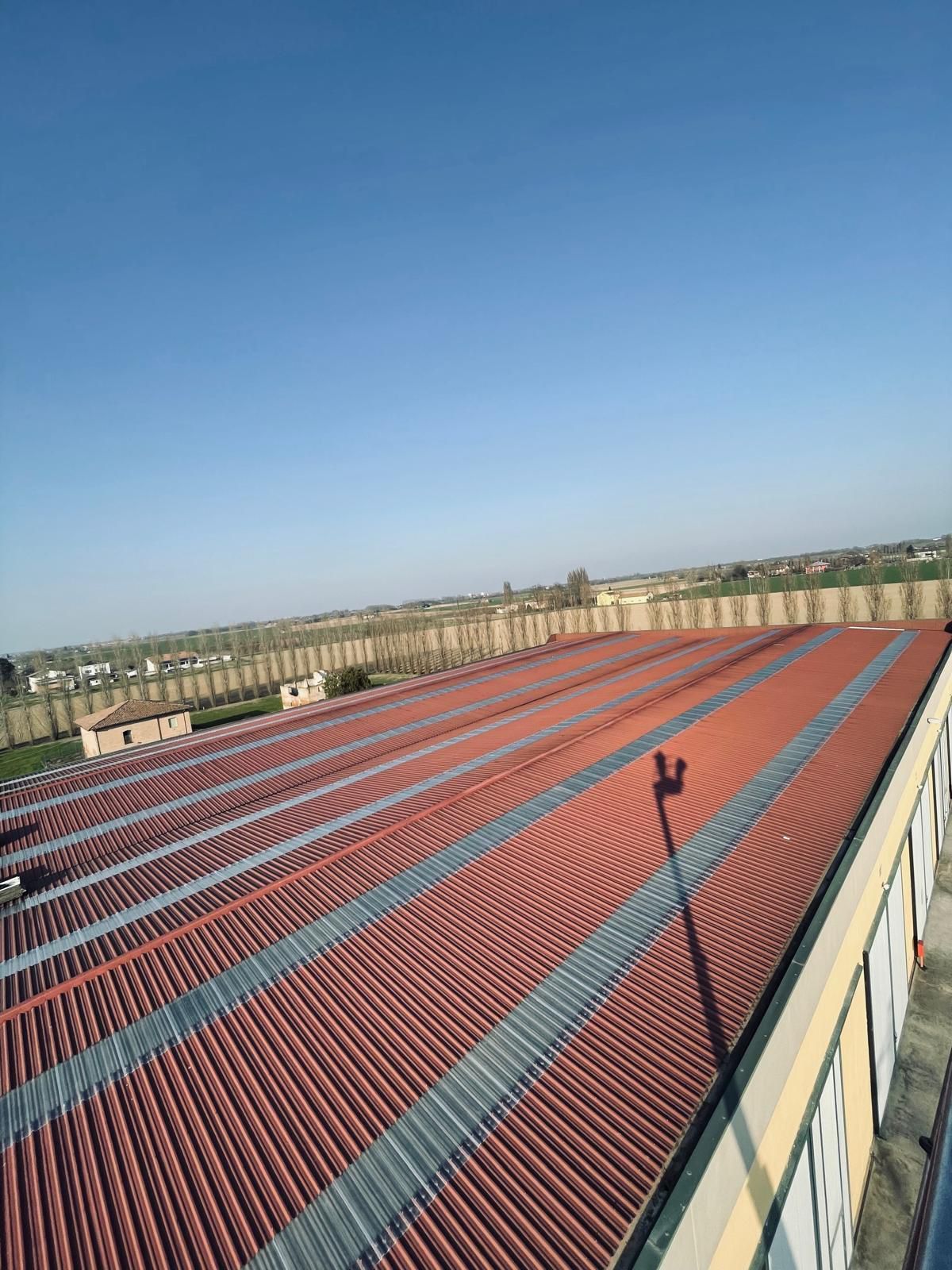 Vista di un tetto di tegole rosse, con colmi di metallo argentato, sotto un cielo azzurro e una recinzione in lontananza.