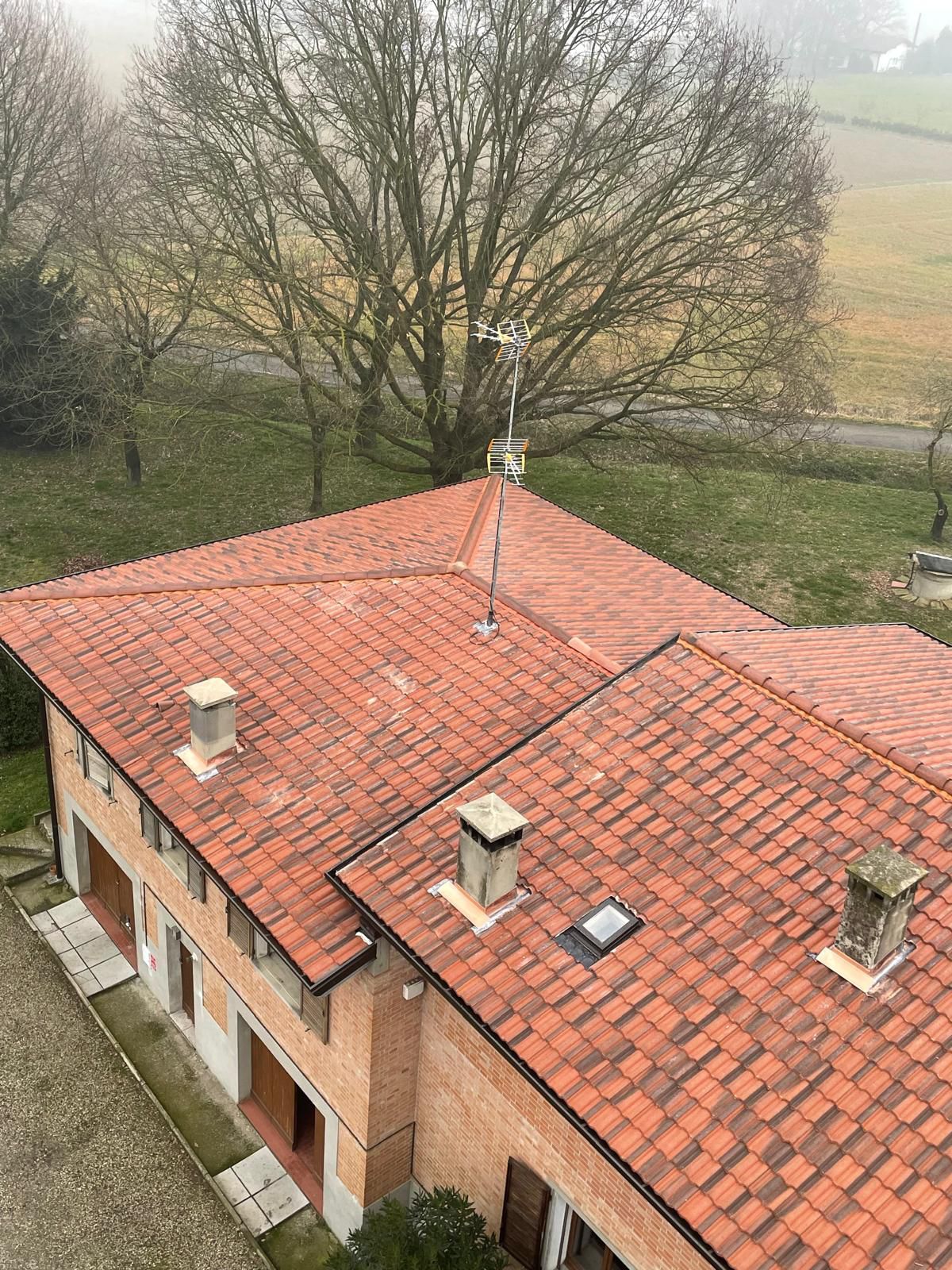 Tetto di tegole rosse di un edificio in mattoni con camini, antenna e vista su un albero e un campo.