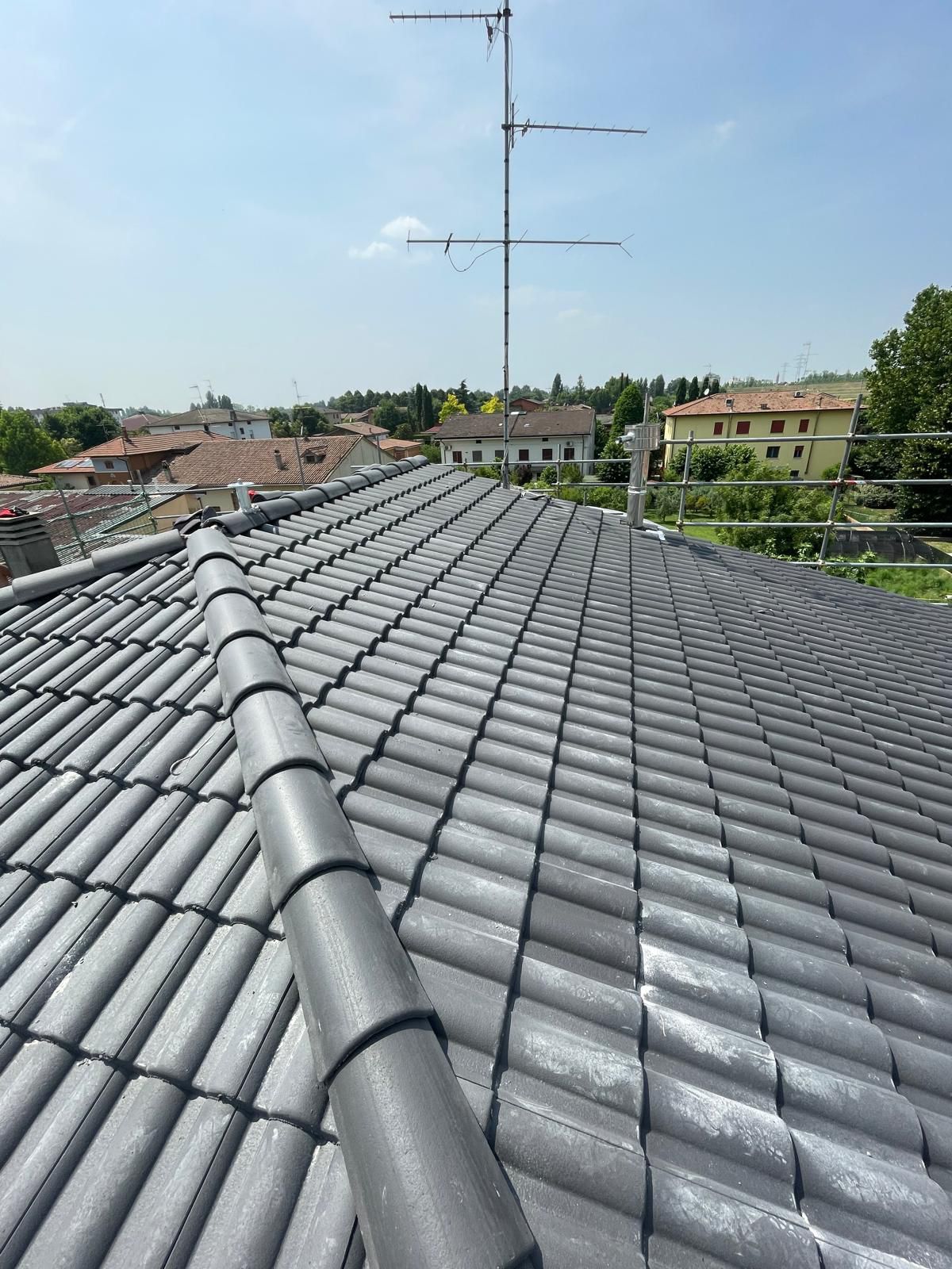 Tetto rivestito di tegole grigie con antenna TV, con vista sul paesaggio urbano sotto un cielo azzurro.
