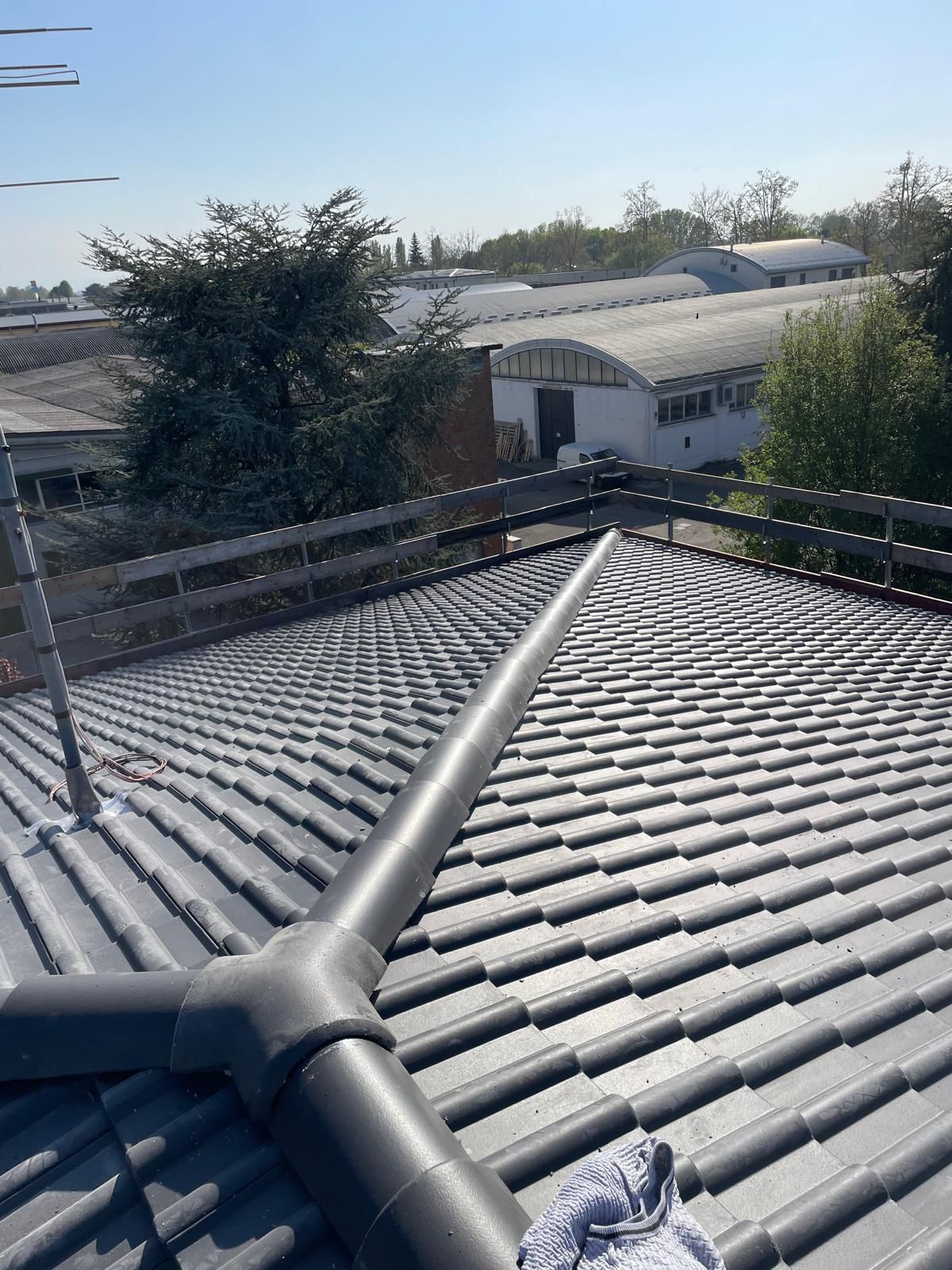 Tetto di tegole grigie con tubi neri e vista di edifici e alberi sotto un cielo azzurro.