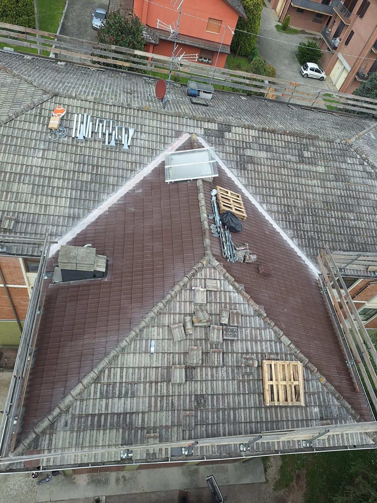 Vista dall'alto di un tetto di tegole rosse con attrezzi e un pallet di legno, con edifici e una strada visibili sullo sfondo.