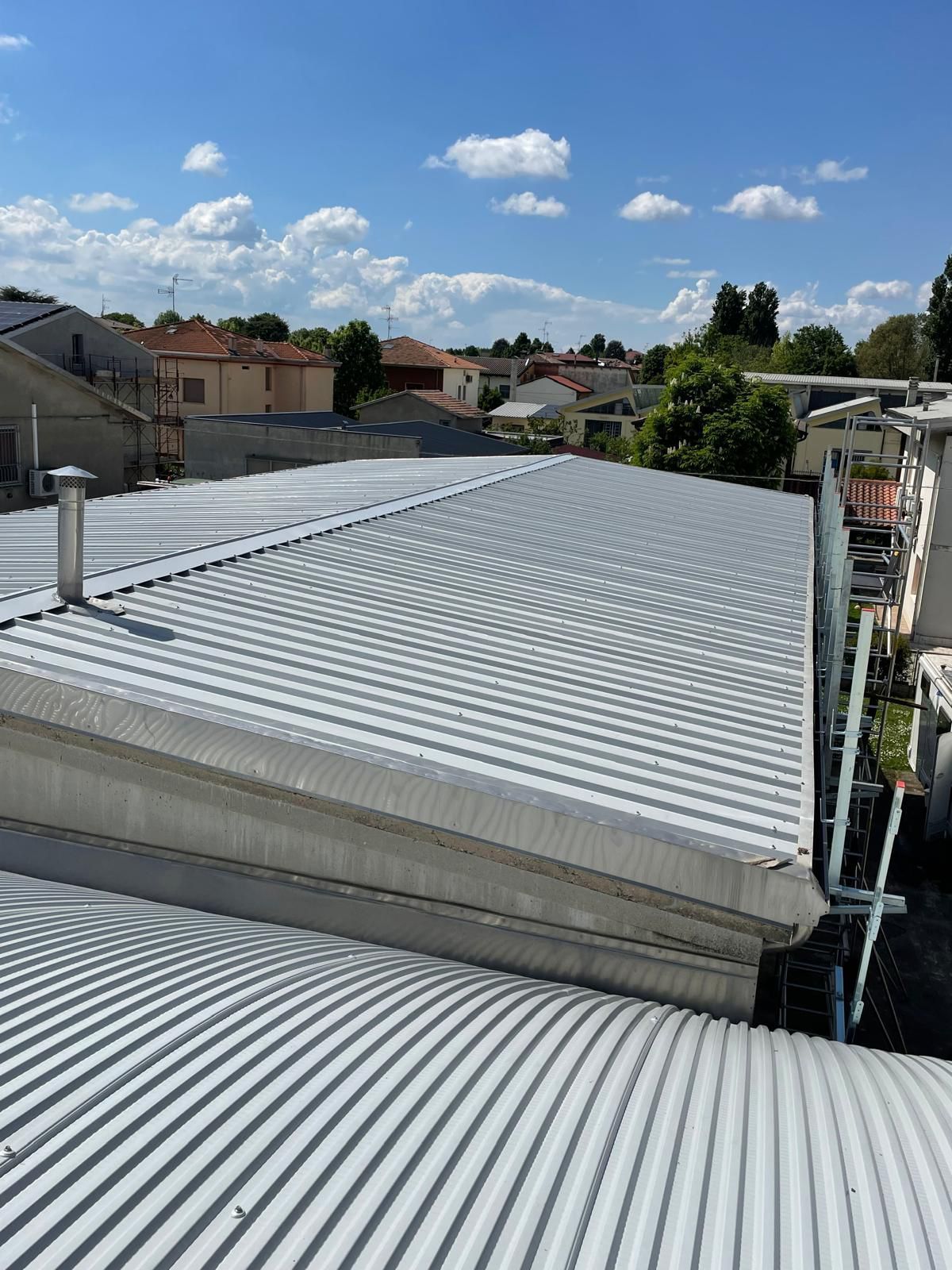 Tetto in lamiera ondulata di un edificio con camino, cielo azzurro e case circostanti sullo sfondo.