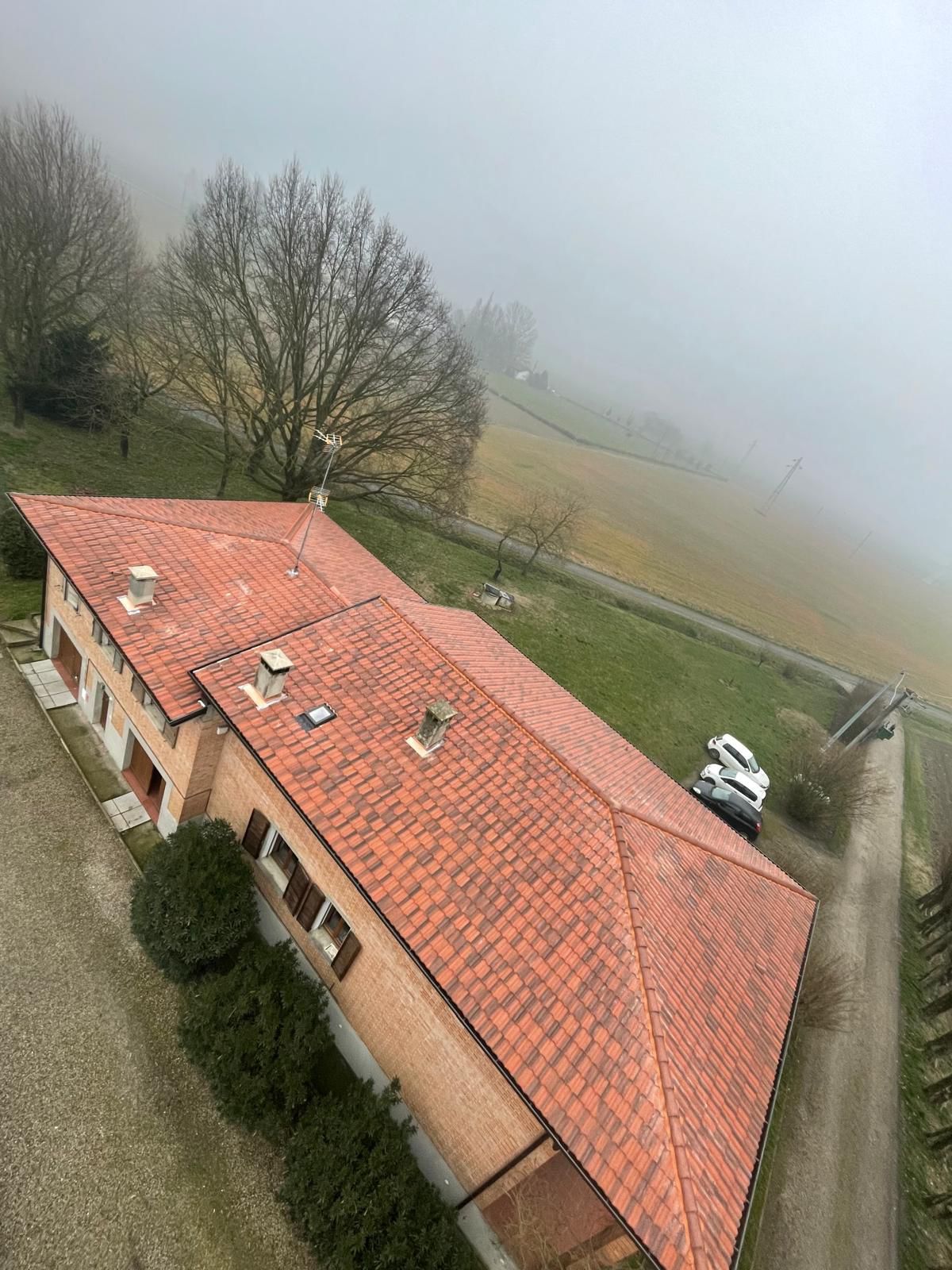 Tetto di tegole rosse di un edificio in mattoni. Sullo sfondo una collina nebbiosa. Strada e veicoli parcheggiati sulla destra.