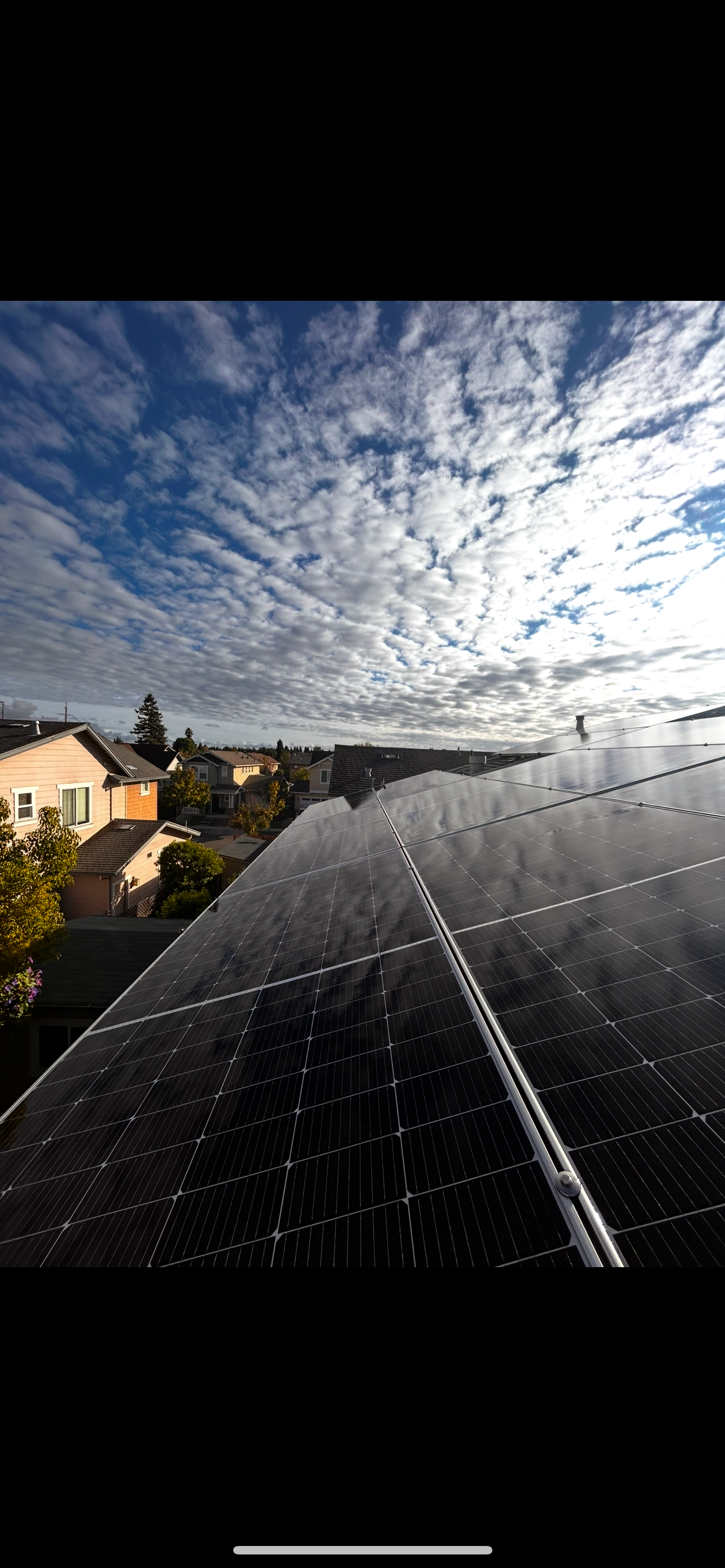 Solar panels on a roof with a cloudy blue sky and houses in the background.