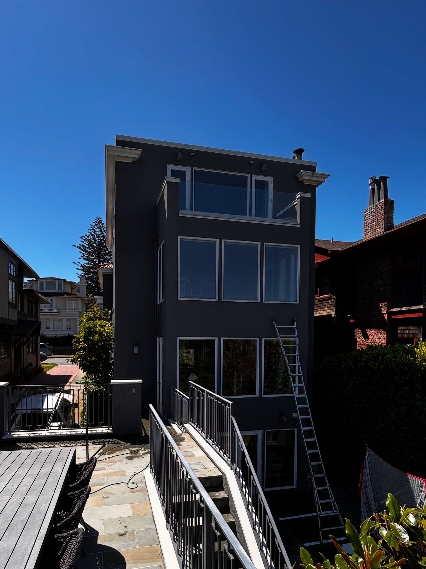 Three-story gray house with large windows, ladder, and access stairs against blue sky.