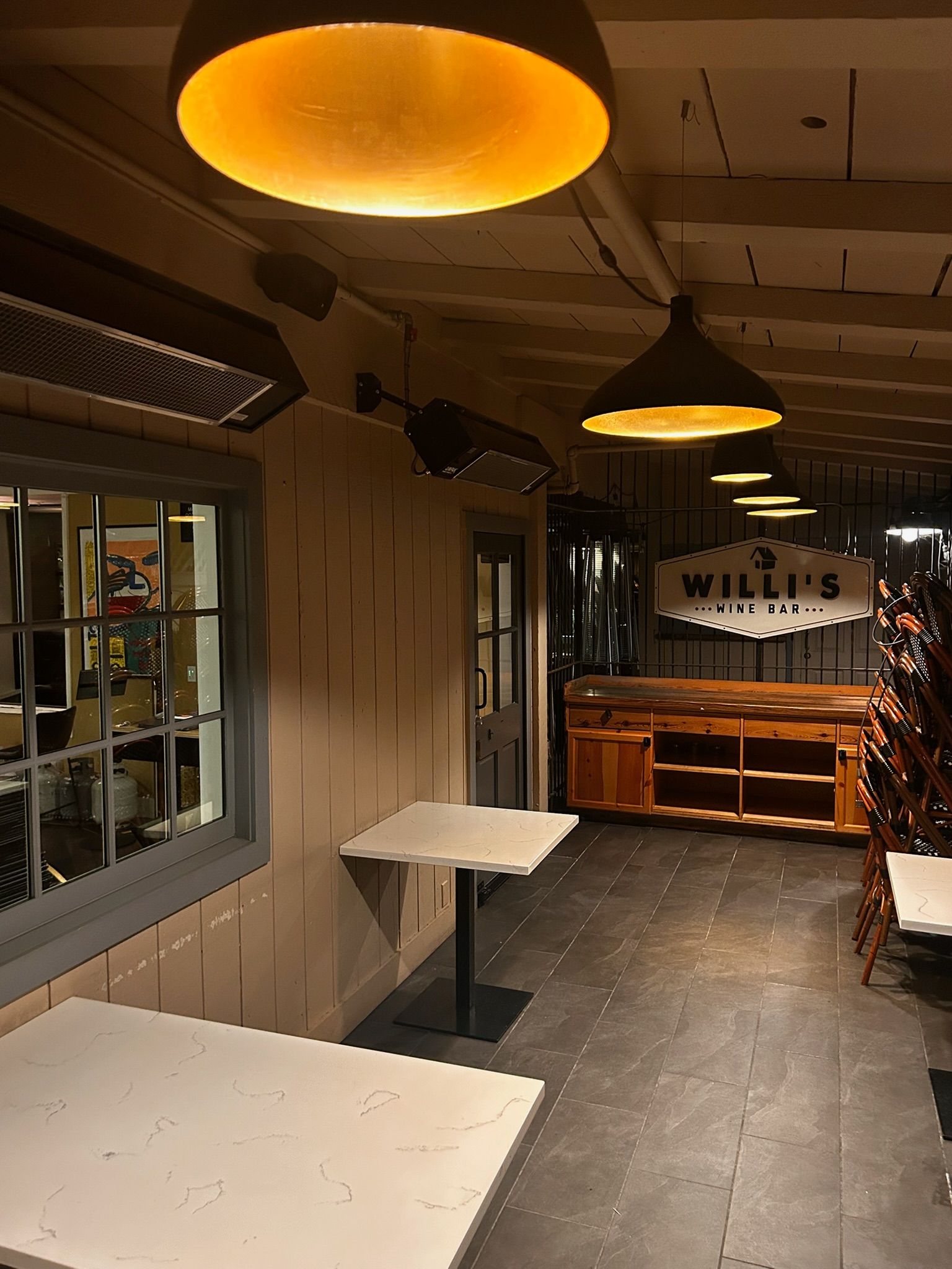 Outdoor restaurant patio with tables, overhead lights, and bar sign that reads 