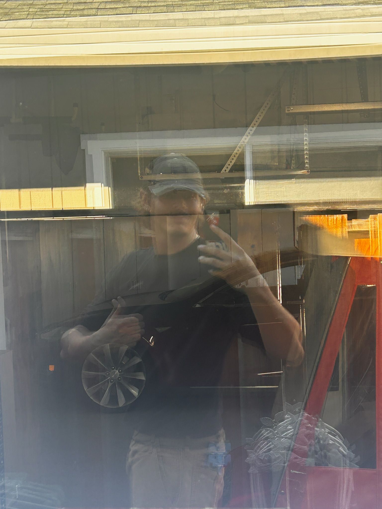 Person reflected in a window holding a circular object, standing outside near a wooden fence and an orange structure.
