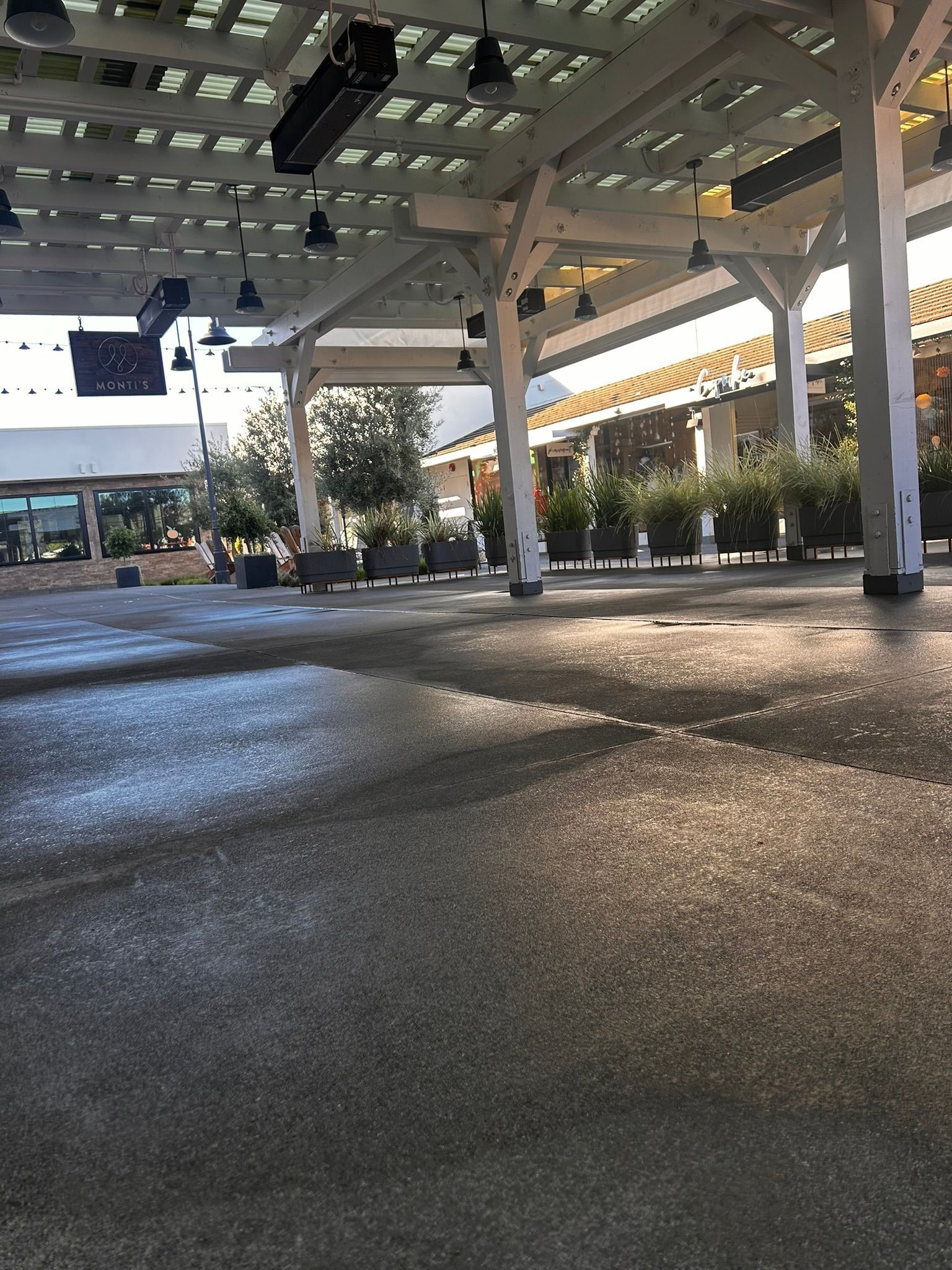 Open-air shopping area with white columns, overhead beams, and rows of potted plants.