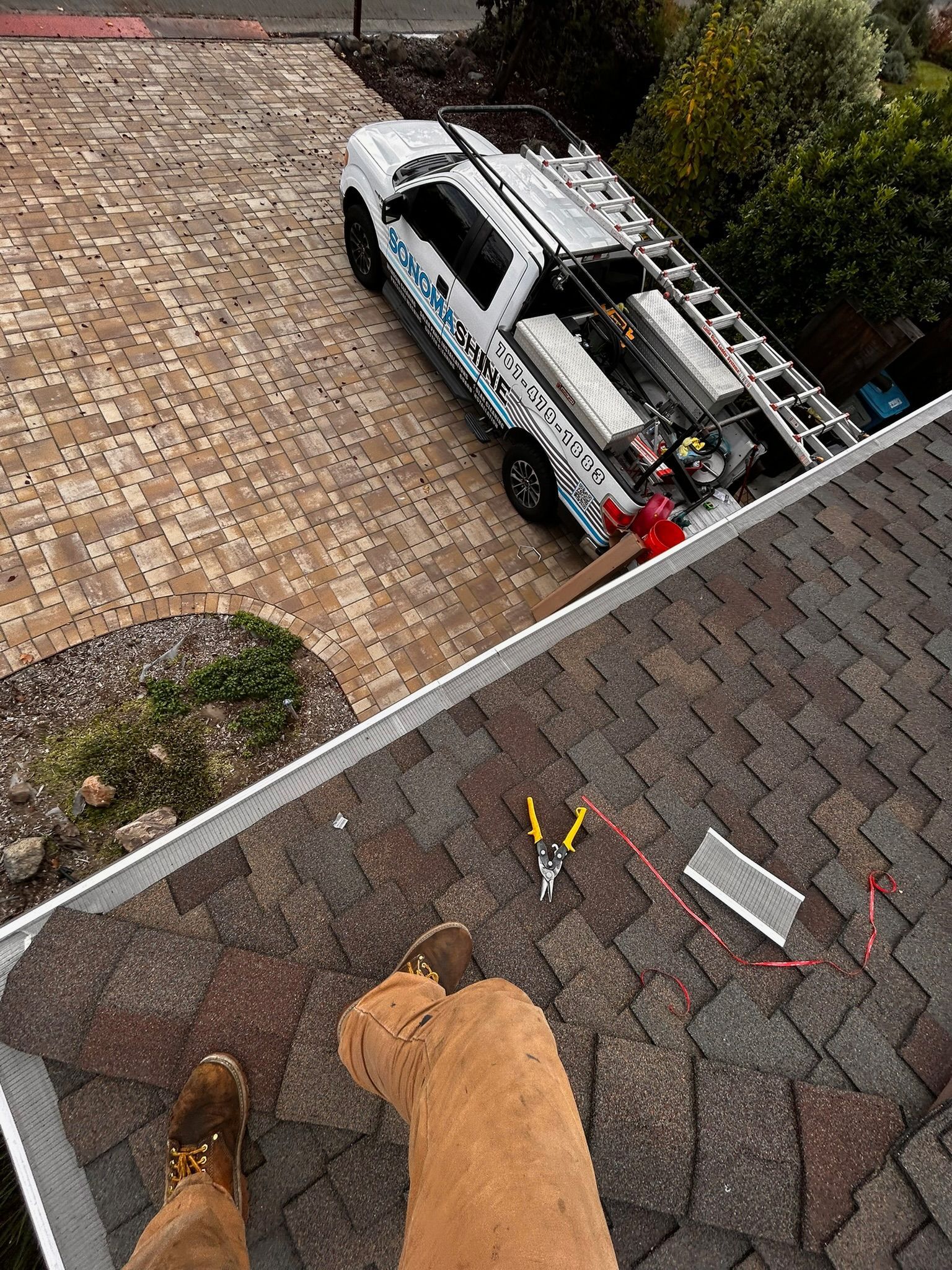 A person on a roof with tools, looking at a truck with ladders parked on a brick driveway.