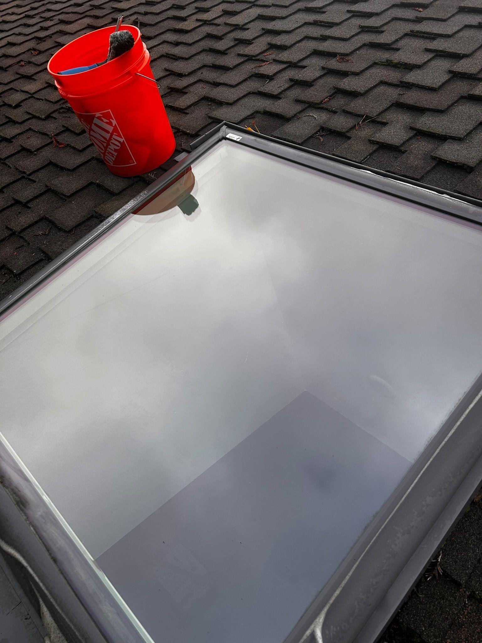 Red bucket on a dark shingled roof next to a skylight reflecting a cloudy sky.