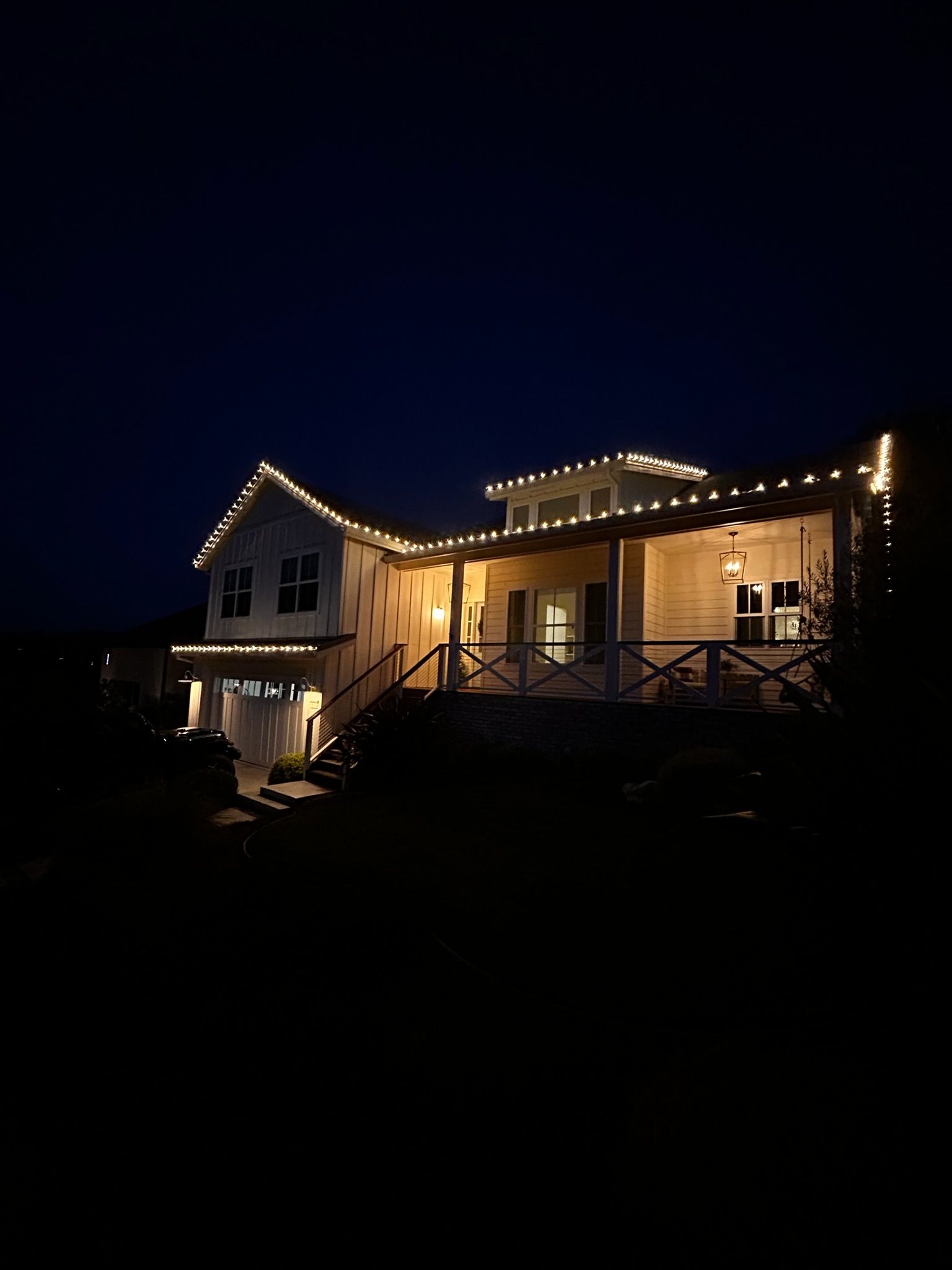 White house at night, lit with string lights along the roof and porch, against a dark blue sky.