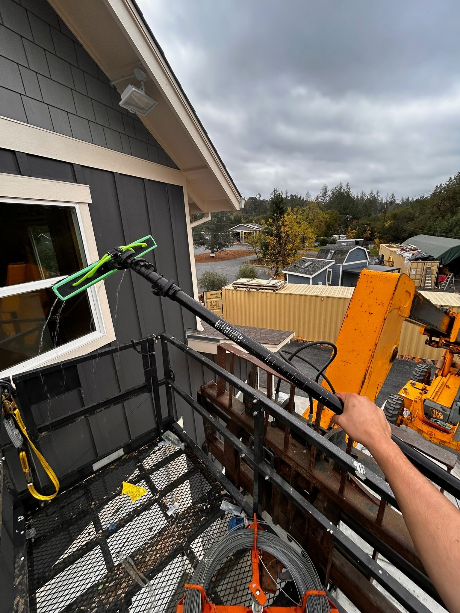 Person washing window on lift, gray house, green brush, cloudy sky.