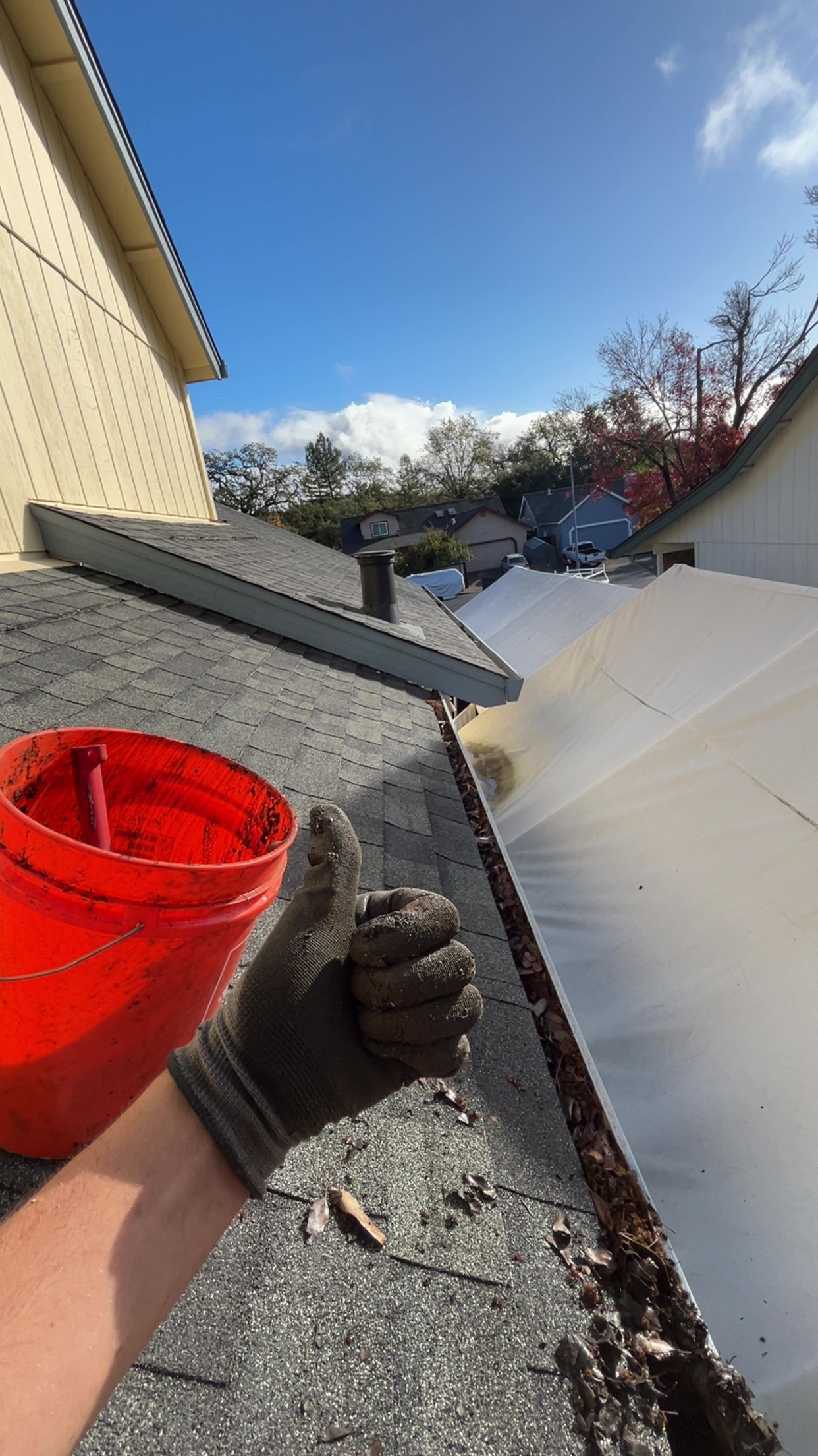 Person in work gloves giving a thumbs-up next to a roof gutter with debris. Bright blue sky overhead.