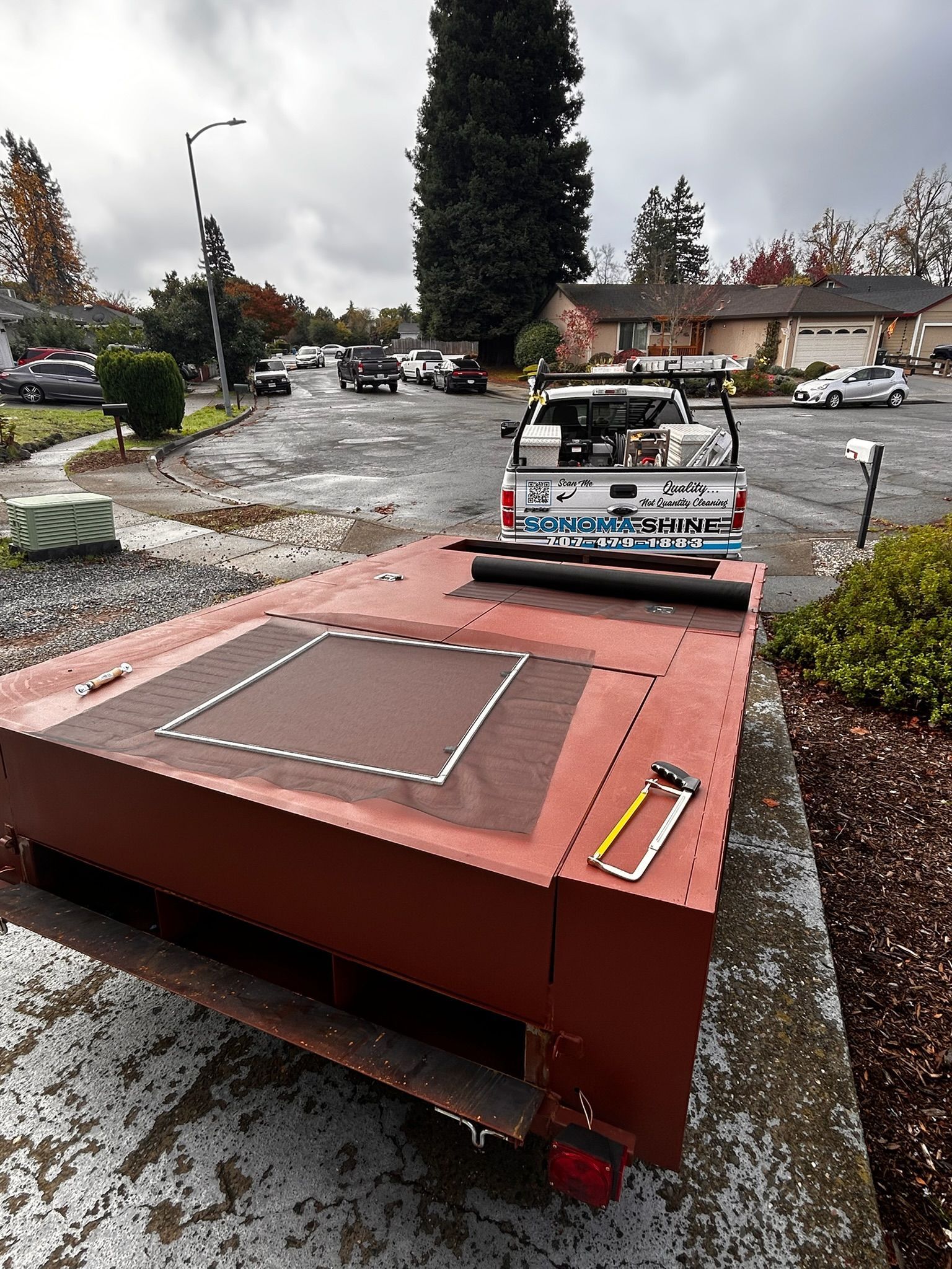 Red trailer parked on a concrete surface with a white pickup truck. Residential street background, cloudy day.