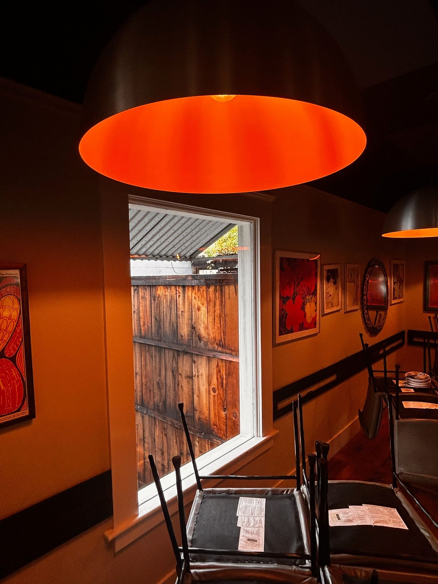 Interior of a restaurant with orange overhead lights, window looking outside onto a wooden fence. 