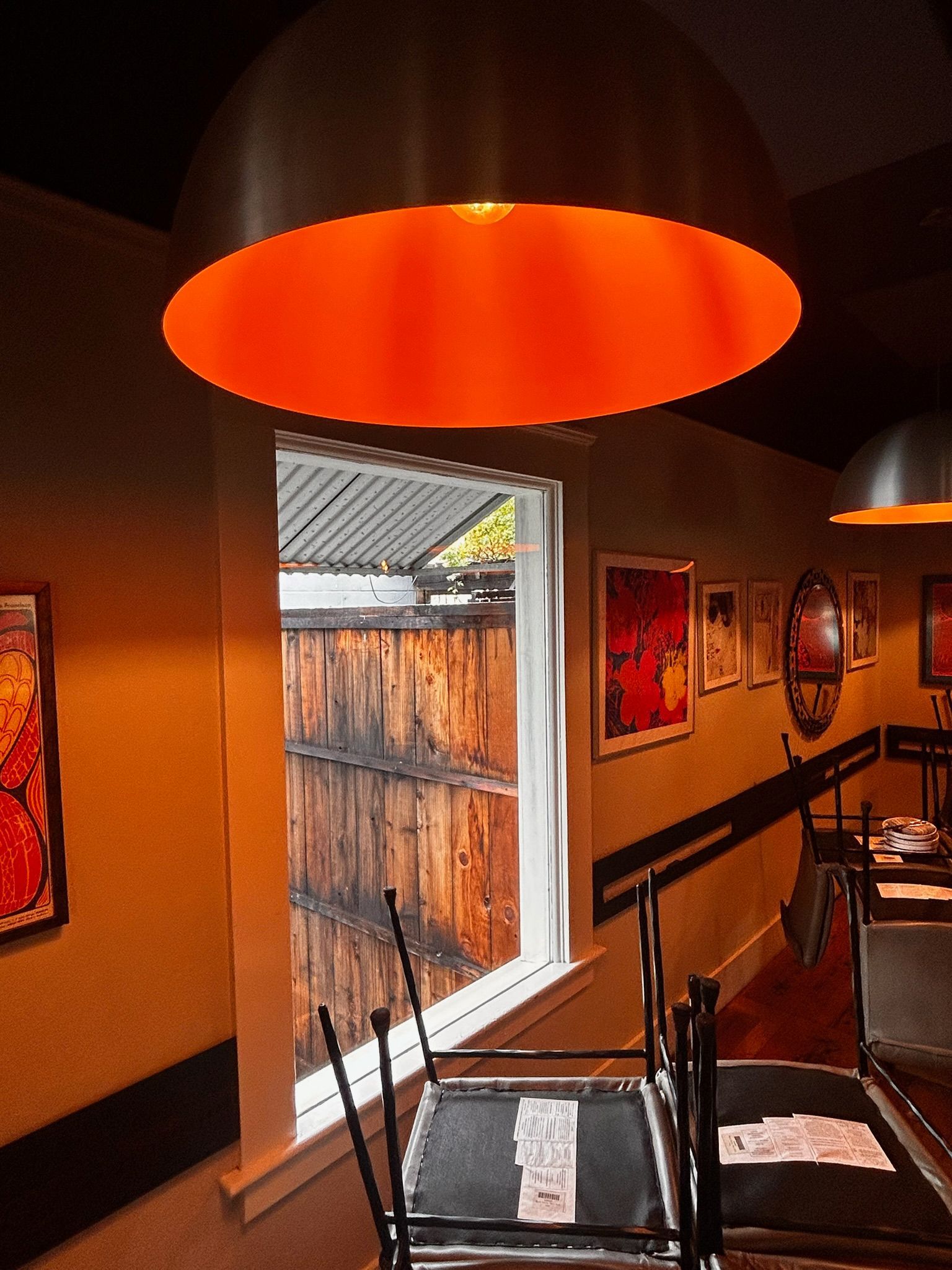 Restaurant interior: orange pendant light, window with wooden fence view, yellow walls, artwork, overturned chair.