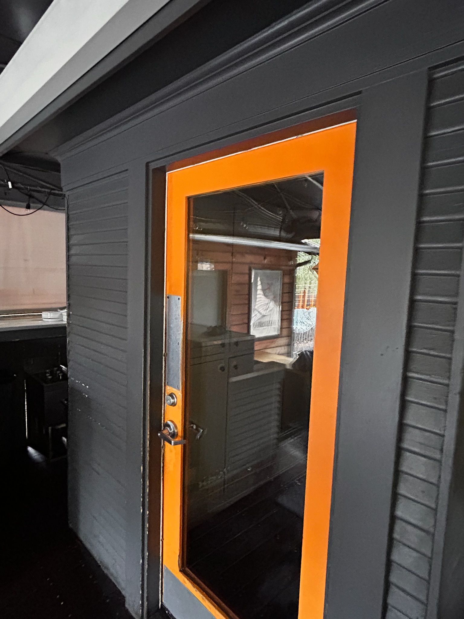 Exterior door with orange frame, glass panel. Dark gray siding. Door handle visible. Interior reflected in the glass.