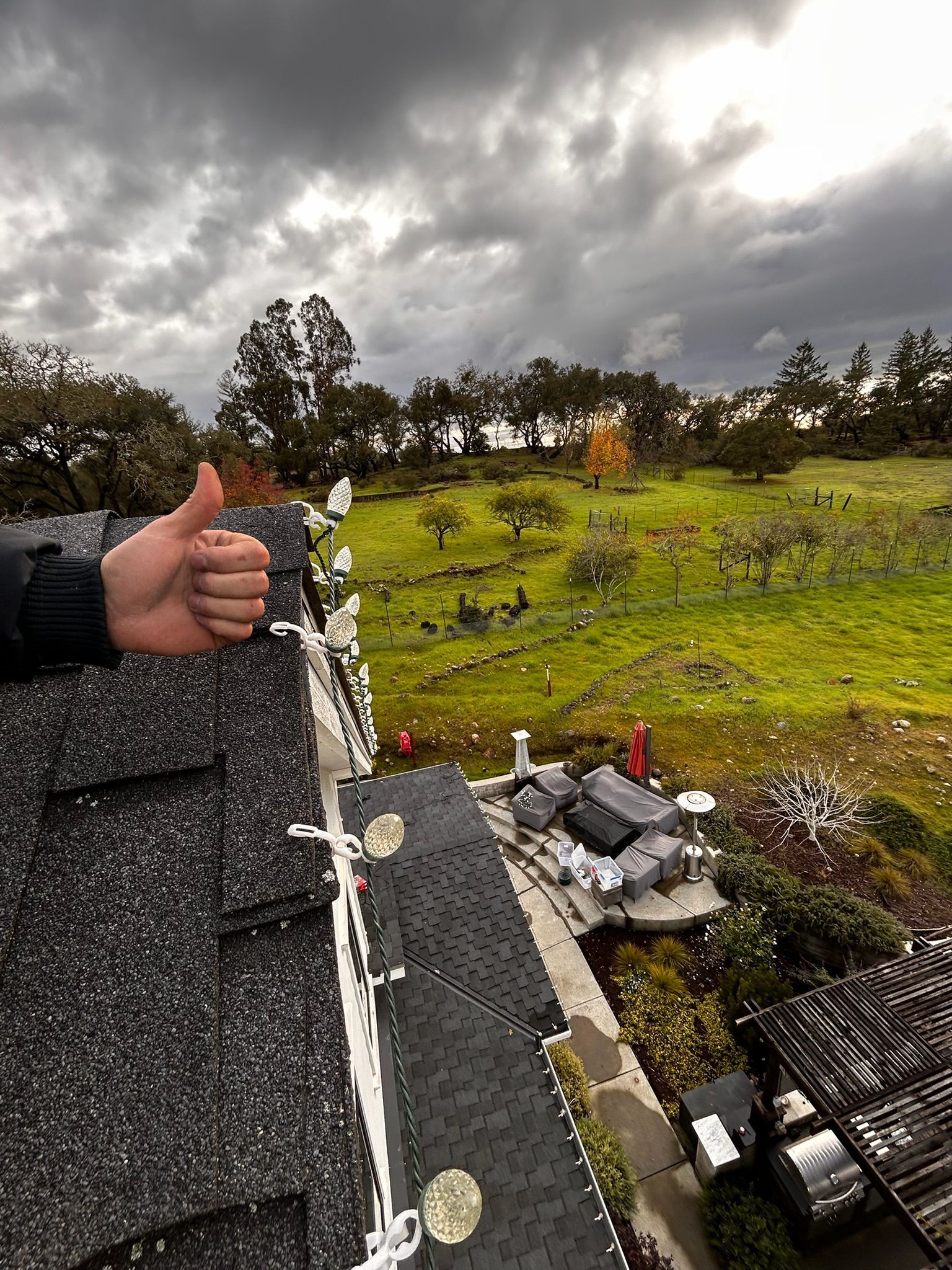 Thumb up from a person on a rooftop with Christmas lights, overlooking a green field and cloudy sky.