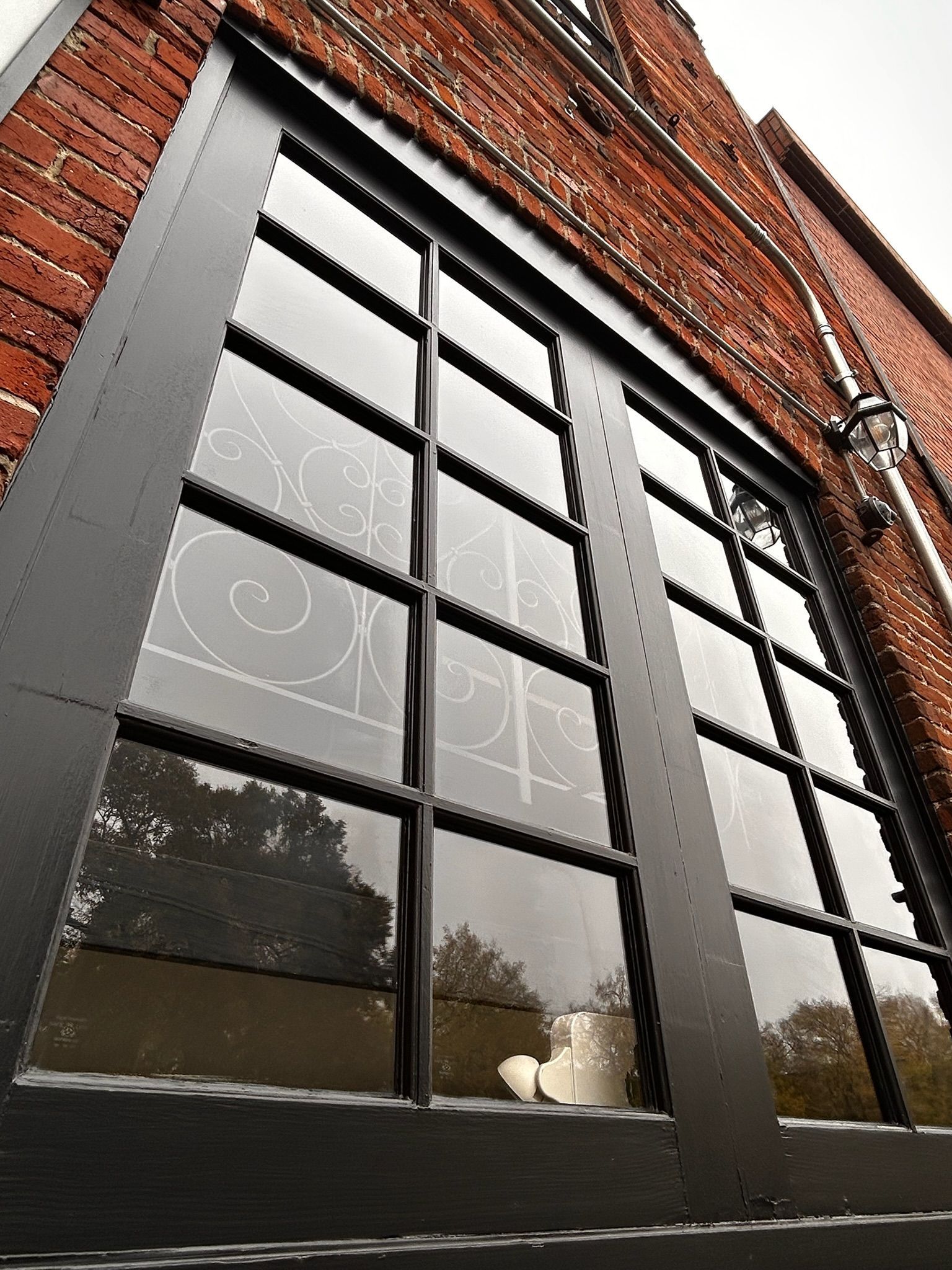 Black-framed window with multiple panes against a red brick building; trees reflected in the glass.