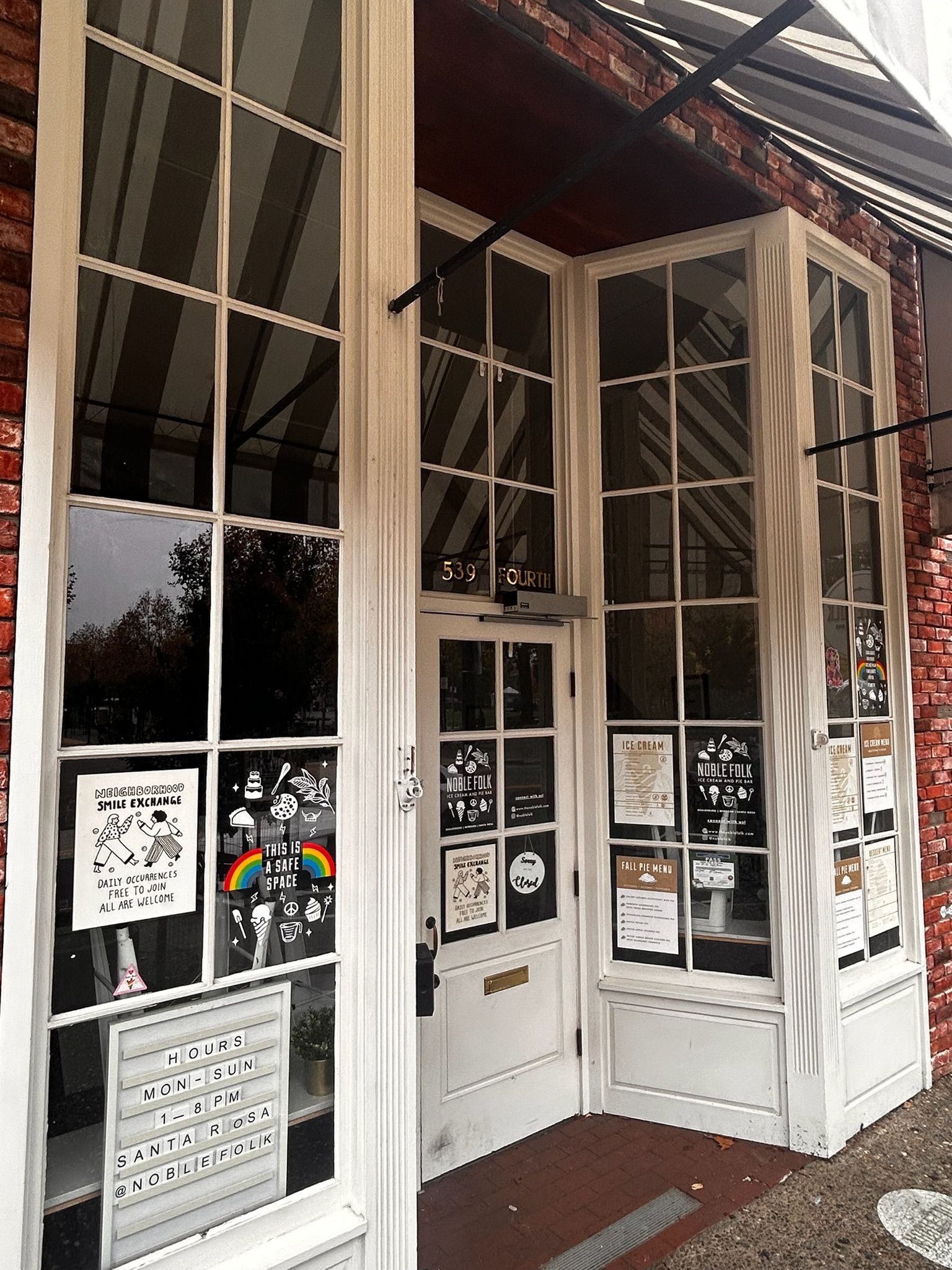 White-framed storefront entrance with large windows; door is ajar. Red brick exterior with awning. Posters in windows.